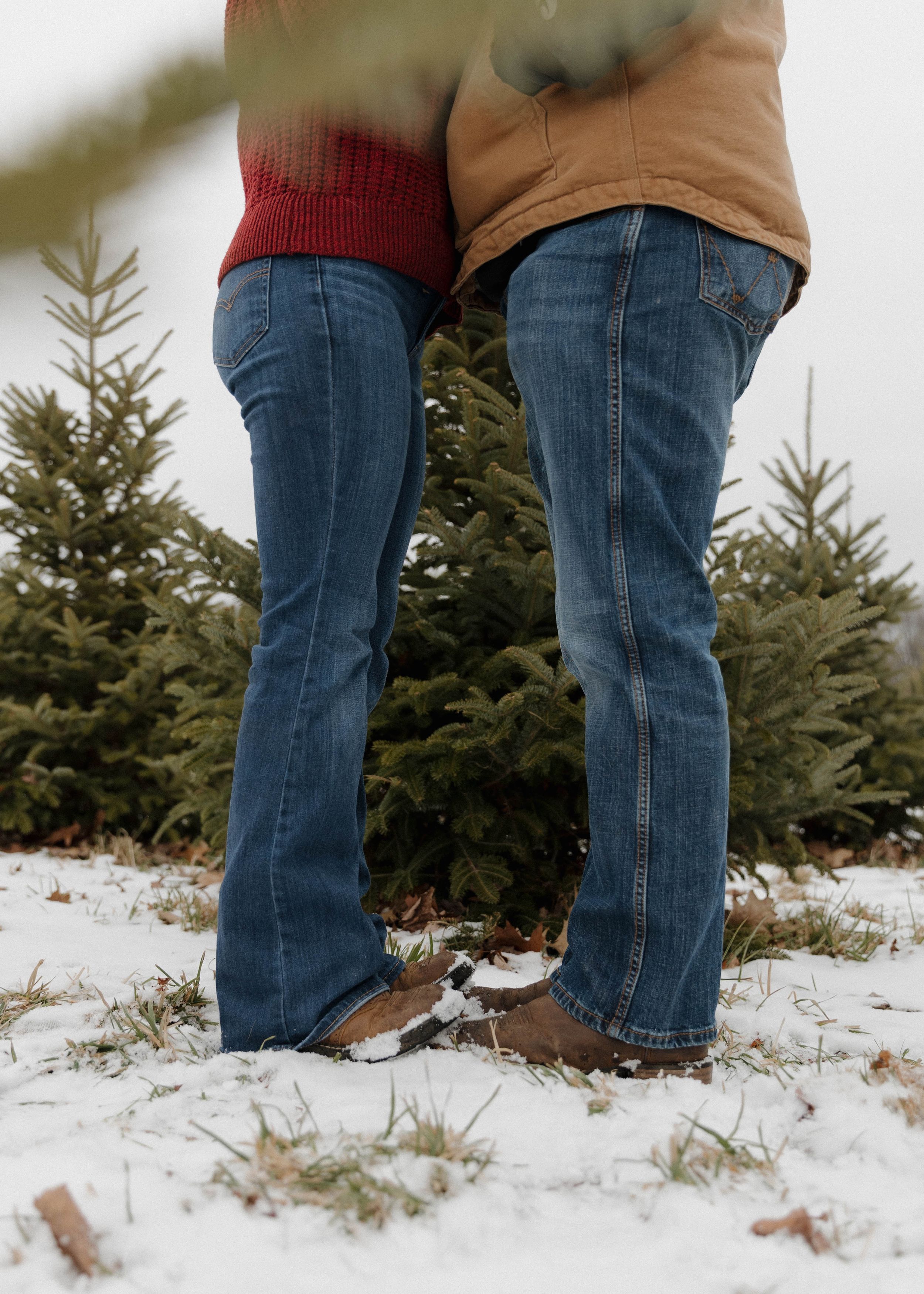 Two people in jeans and winter boots stand in snowy ground near evergreen trees at a Christmas tree farm.
