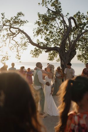 Couple shares first dance at golden hour beach wedding ceremony under a twisted tree with guests surrounding them.