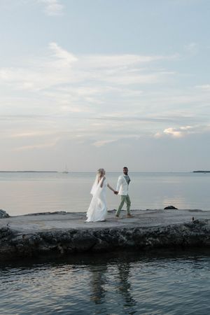 Bride and groom holding hands walking on a stone pier by the ocean at sunset during their beach wedding.