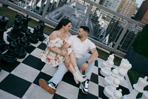 Couple sitting together on a giant outdoor rooftop chess board surrounded by large chess pieces in an urban setting.