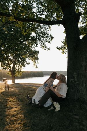 Couple sits beneath autumn trees at sunset casting long shadows across a grassy field.