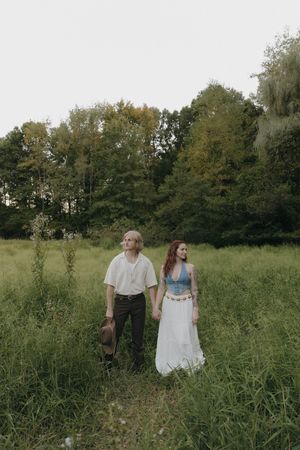 Couple walking hand in hand through tall grass in a meadow with trees in the background on a summer day.