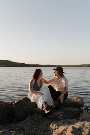 A couple sits together on rocky coastal shore at sunset enjoying views over a tranquil lake.