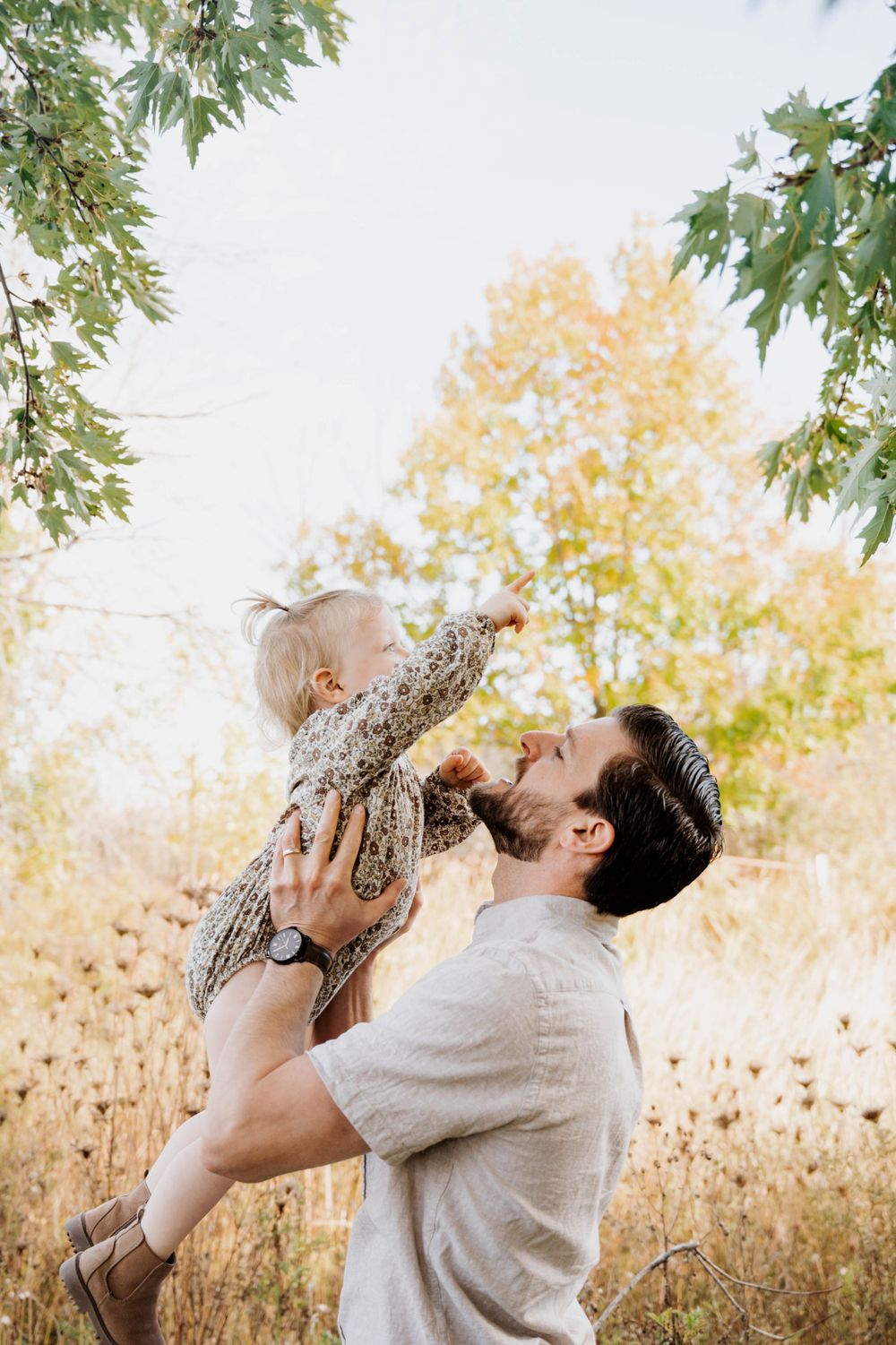 A tender moment captured outdoors as parent lifts small child in floral dress against autumn foliage background.