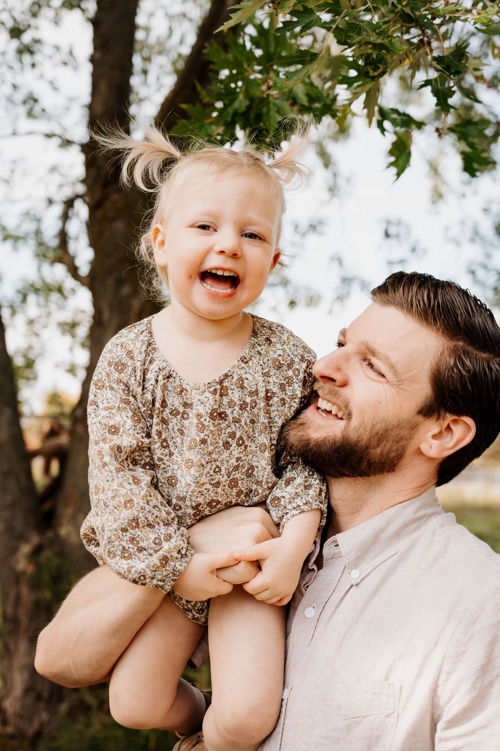 Joyful interaction between parent and toddler beneath tree branches in natural outdoor setting.