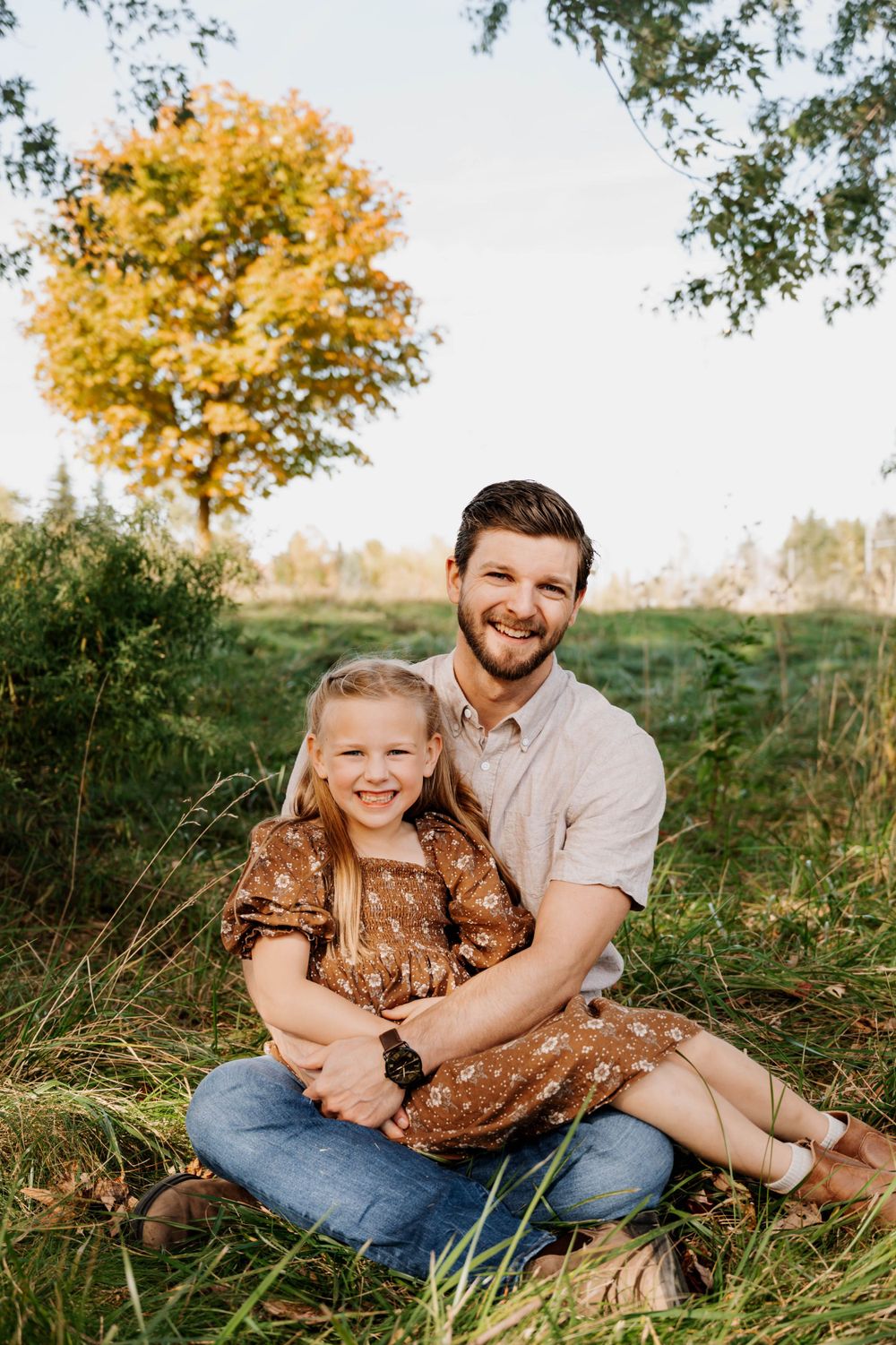 Parent and child sitting together in grass with golden autumn tree in background.