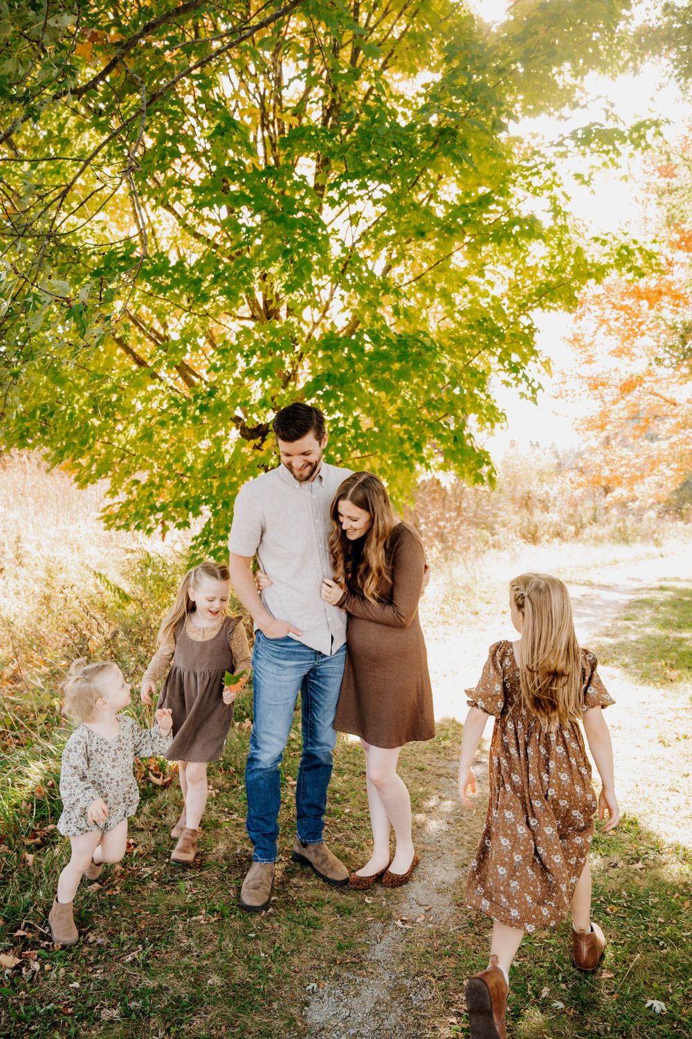 A family walks together on a sunlit autumn path beneath golden trees wearing coordinated brown and neutral tones.