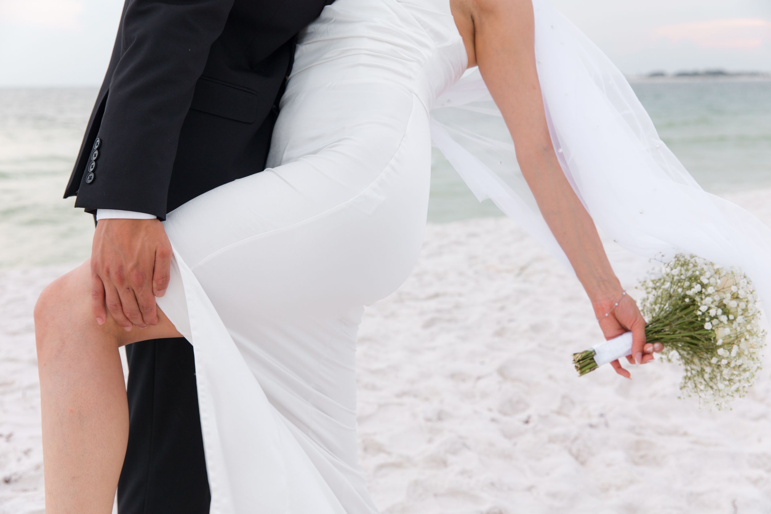 Newlywed couple's legs on beach with bride holding small bouquet.