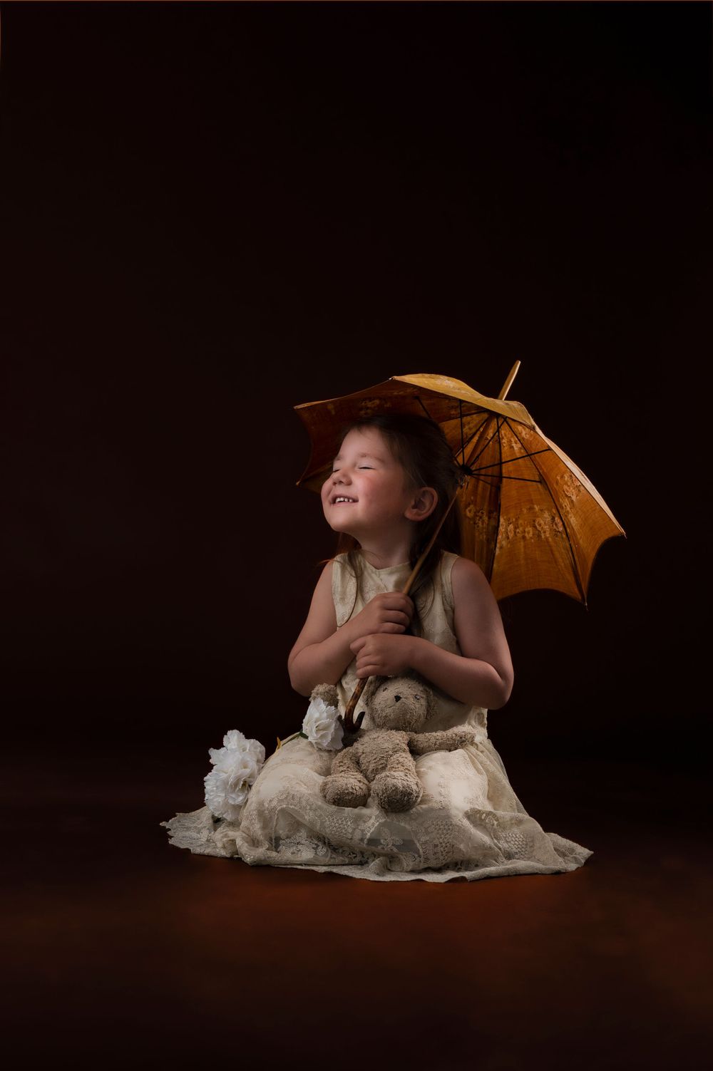 Studio portrait of a small child in a white lace dress holding a vintage parasol against dark background.