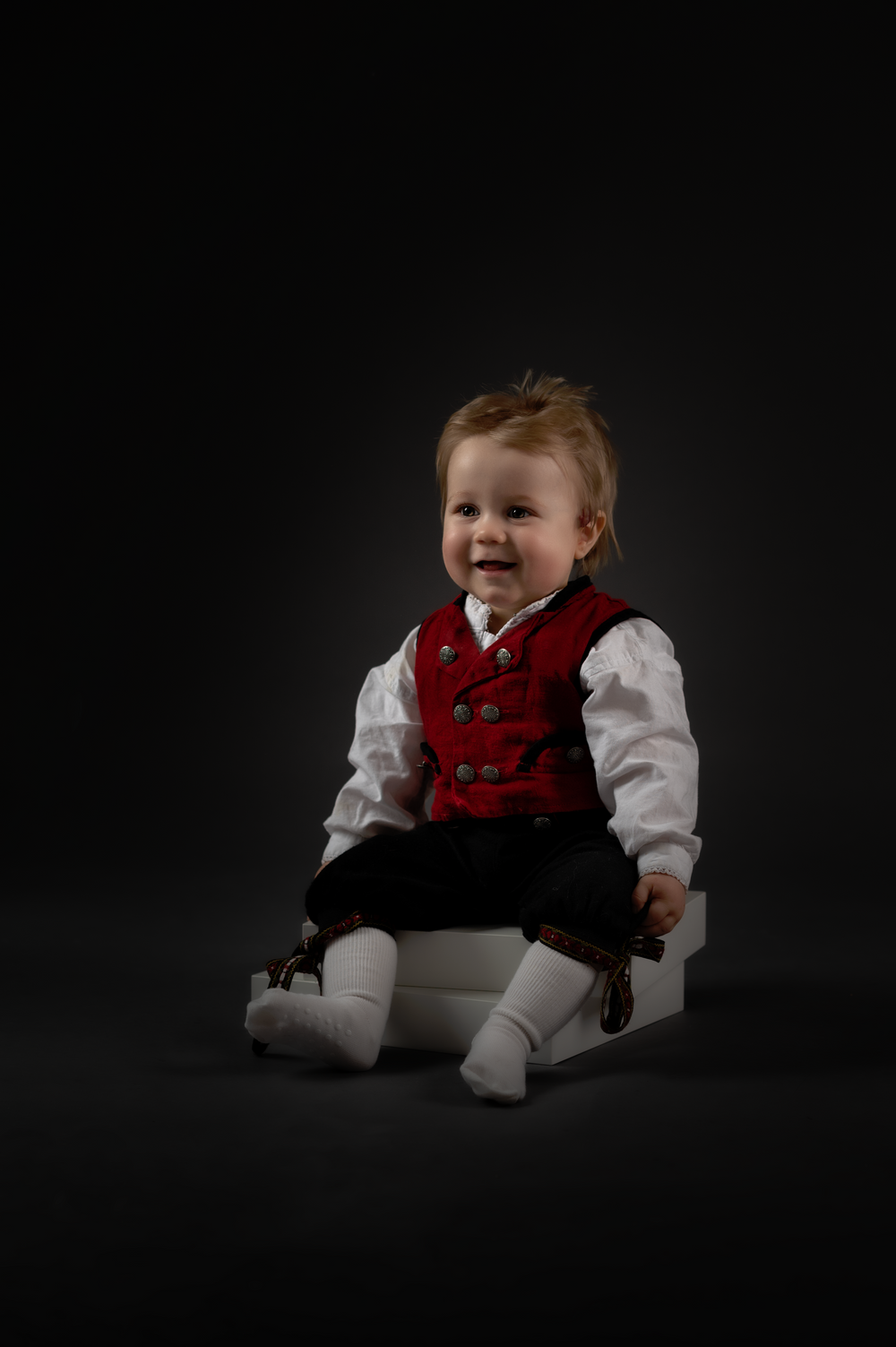Studio portrait of a child wearing formal red vest and white shirt against black background.