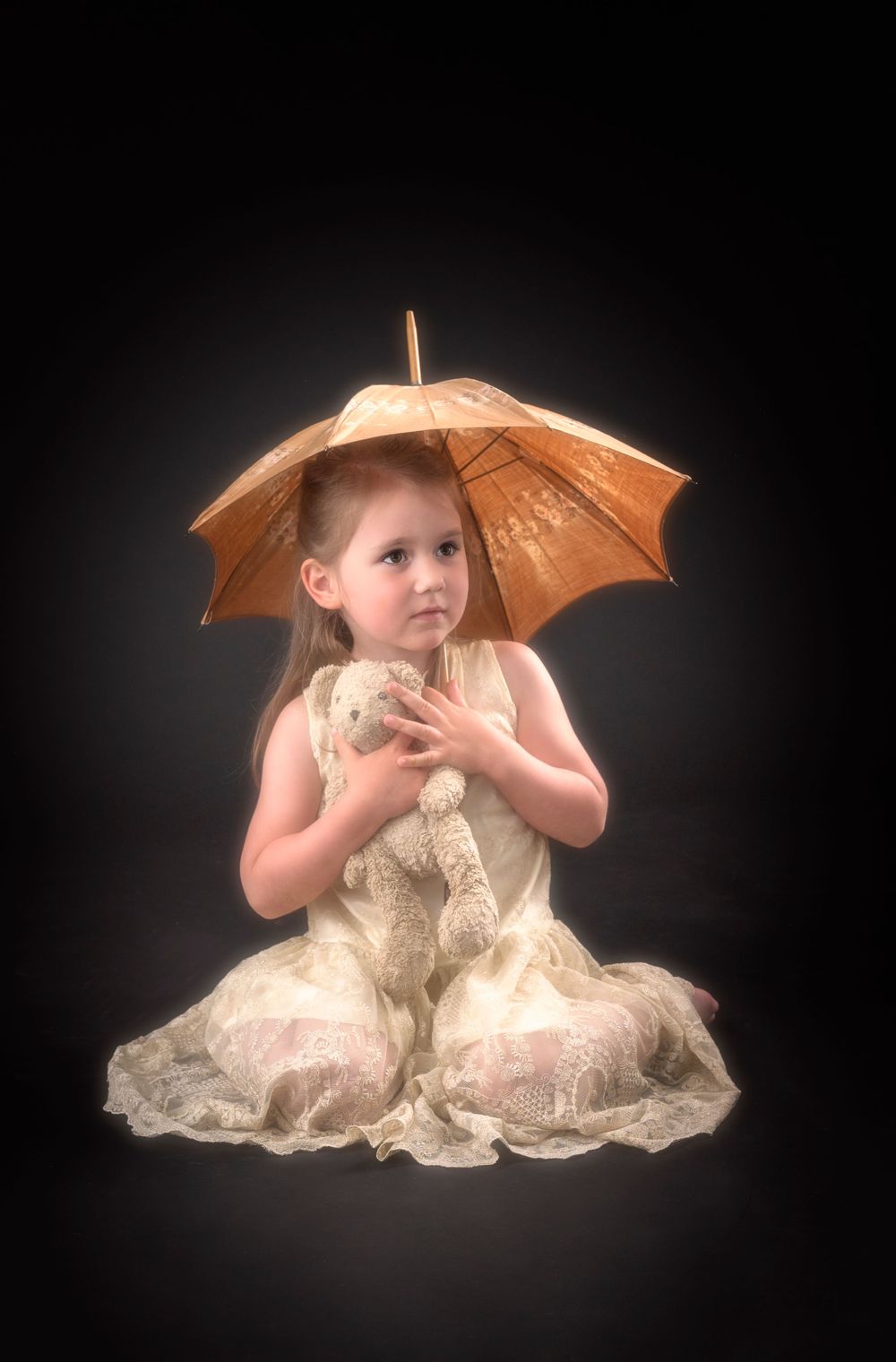 Artistic photo of a child with orange parasol in white lace dress on dark backdrop.