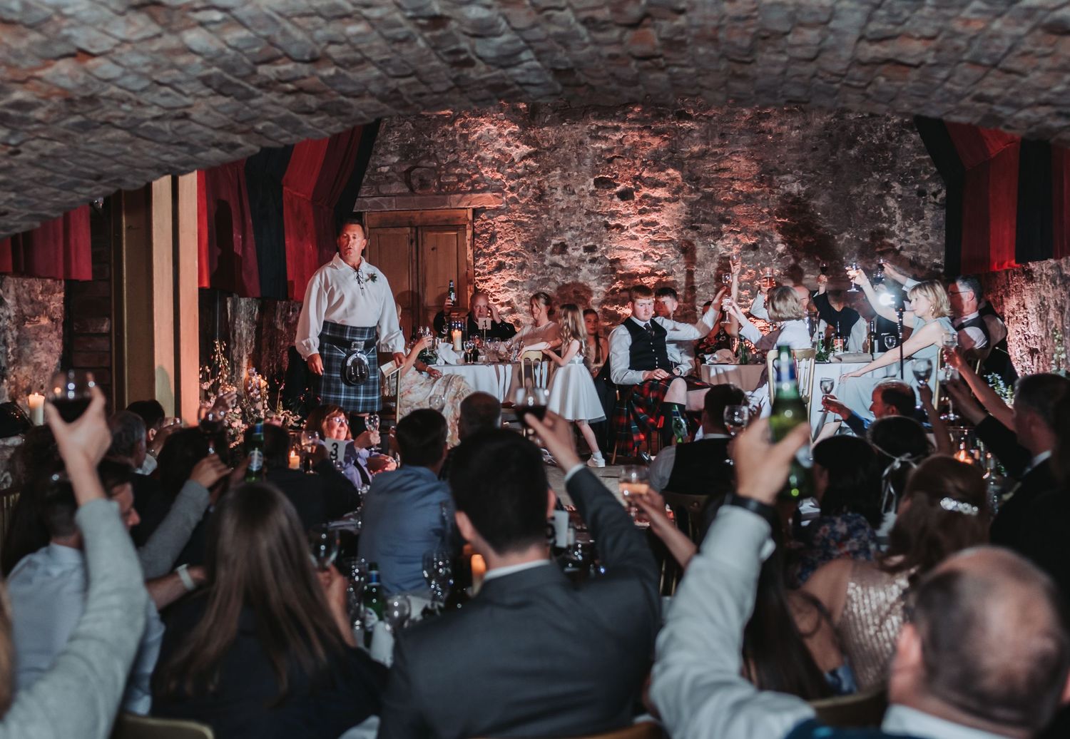 A lively wedding celebration in a rustic cellar venue with guests dancing and raising their hands.