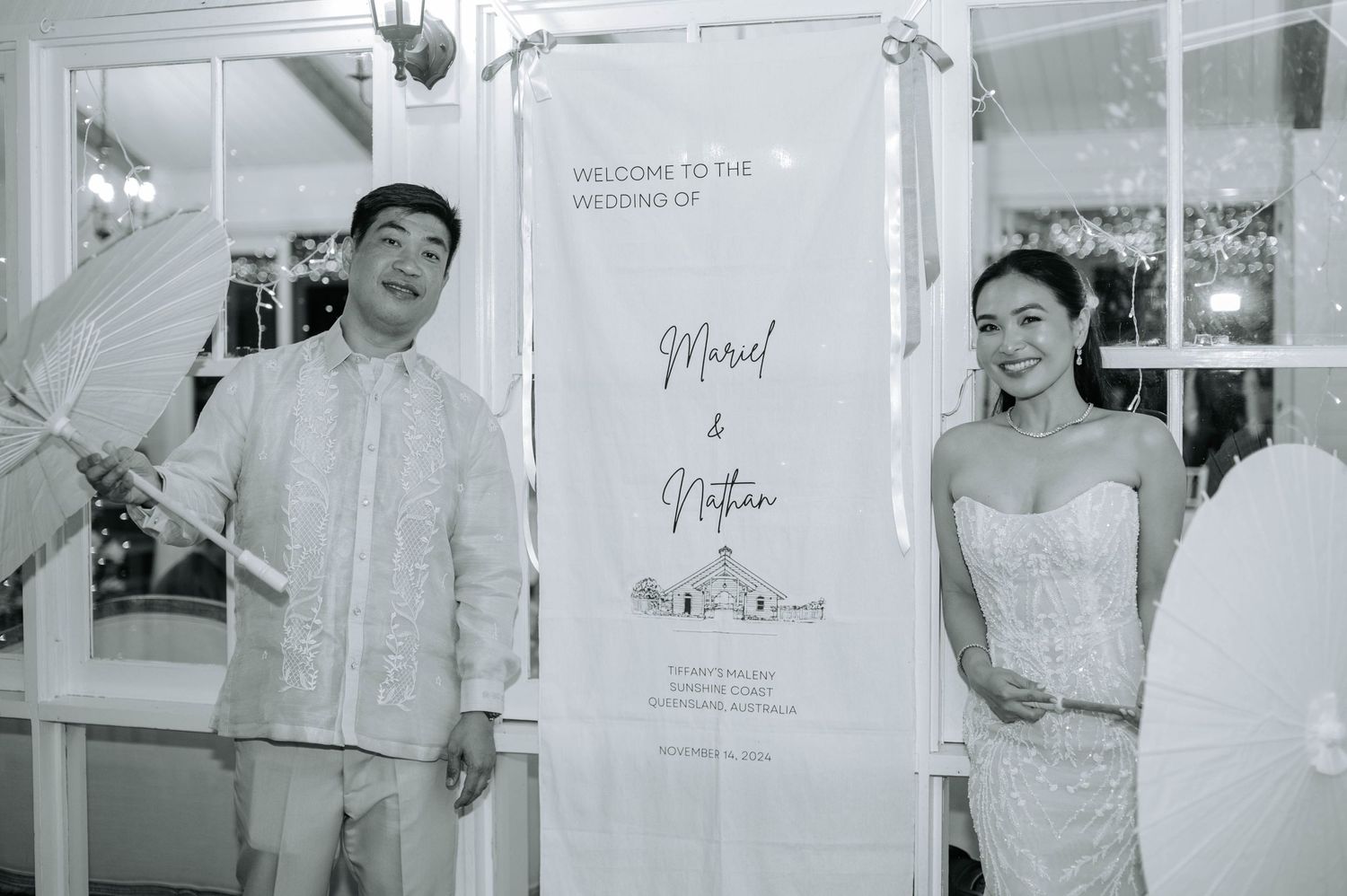Wedding couple in formal attire posing next to their wedding signage in black and white.