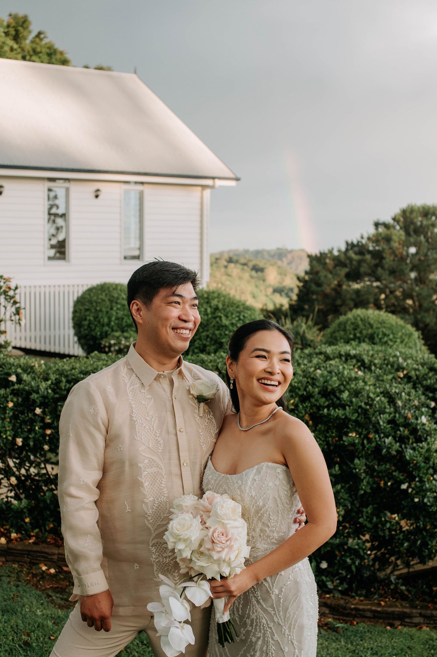 Wedding couple smiling and holding bouquet in front of white house with rainbow in background.