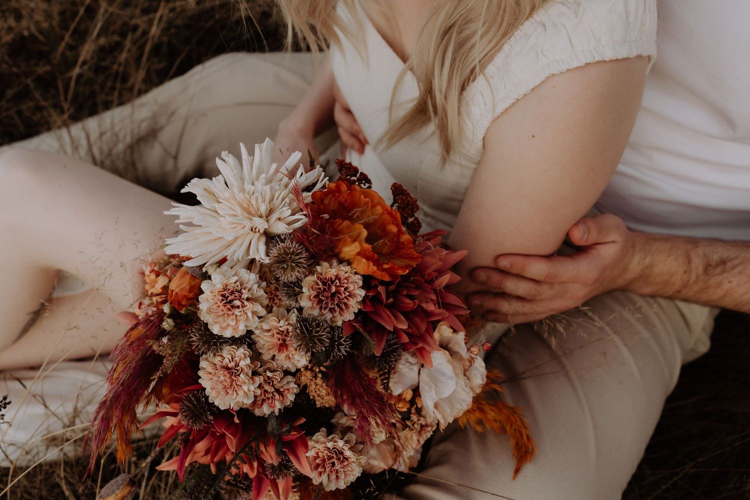 Close-up of autumn-colored wedding bouquet with white dahlias and burgundy leaves on white fabric.
