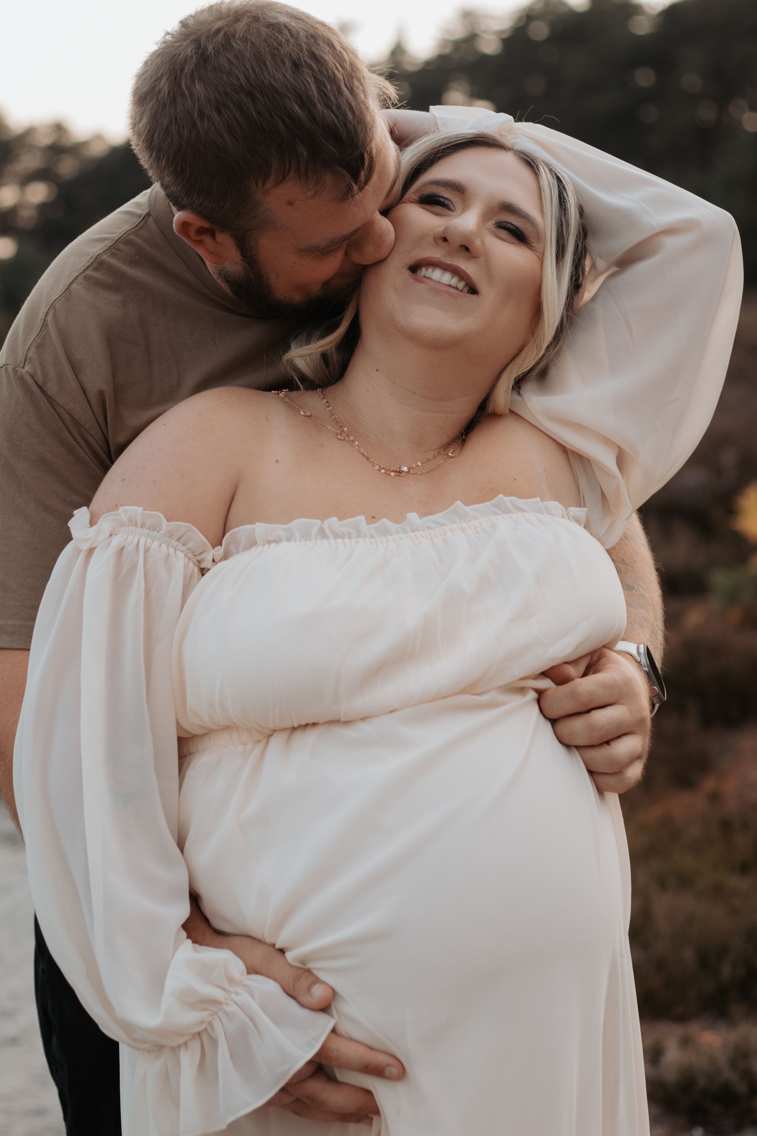 Smiling pregnant woman in white off-shoulder dress embraced by bearded man kissing her cheek outdoors at sunset.