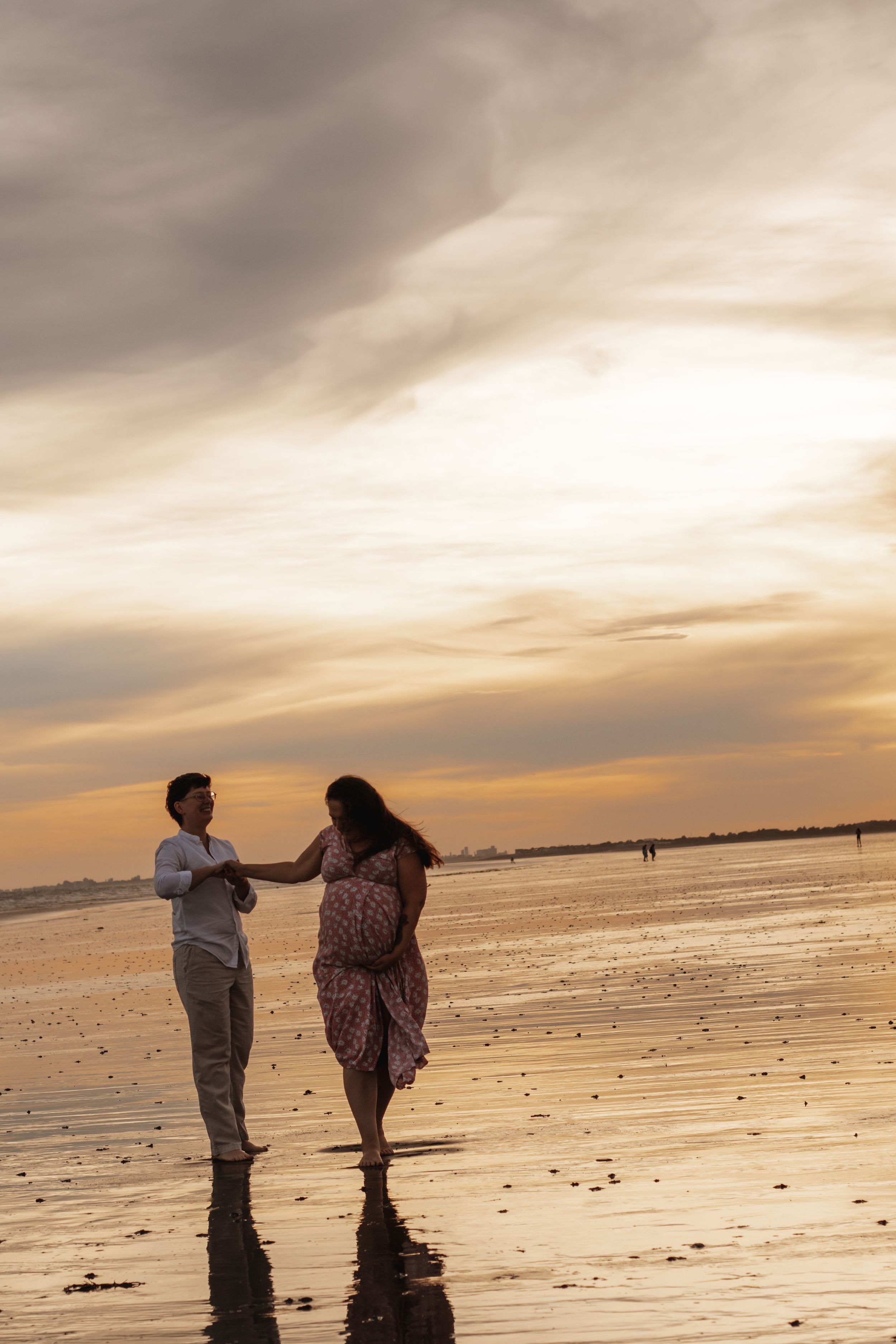 Couple holding hands on a reflective beach at sunset with dramatic golden cloudy sky.