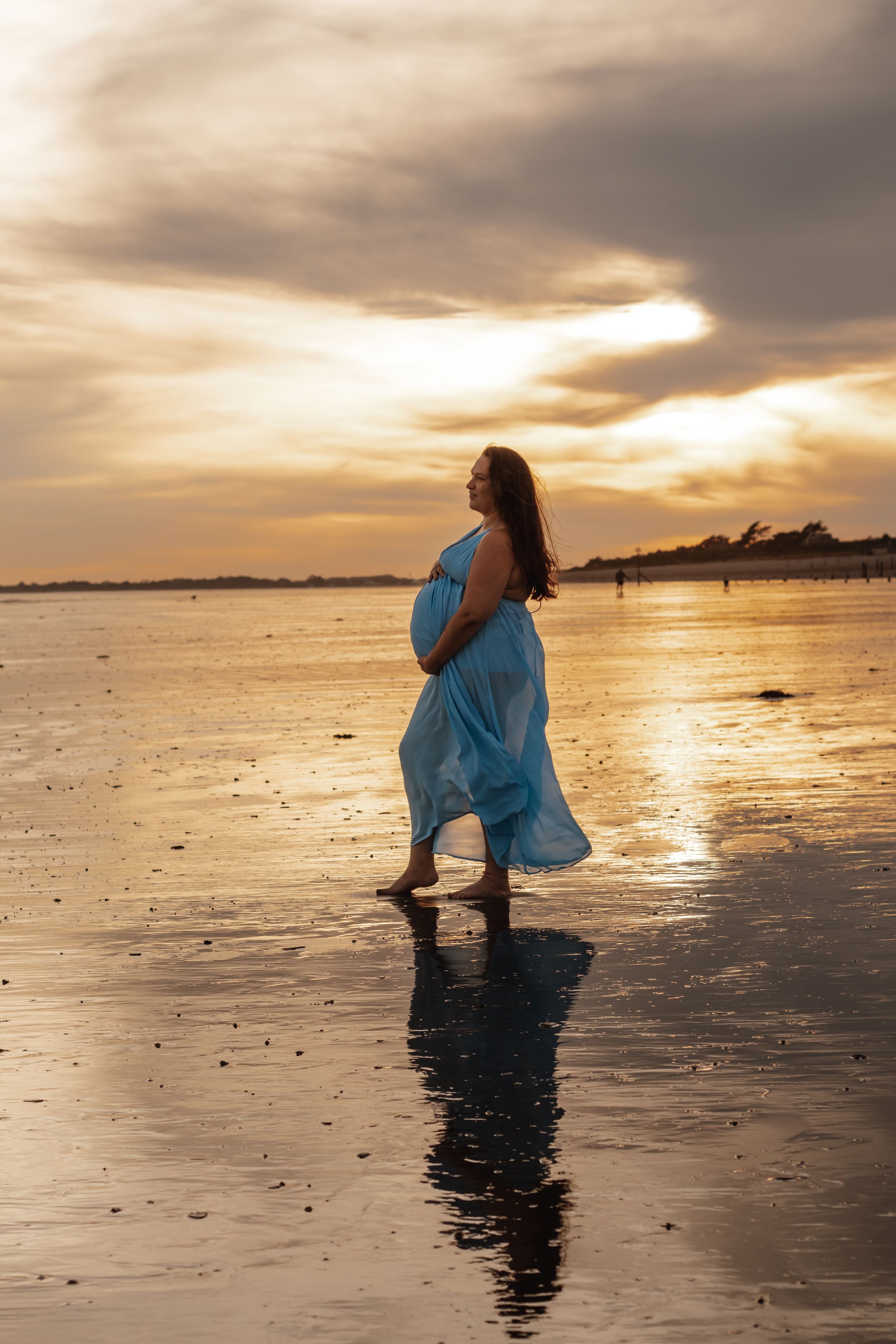 Pregnant woman in flowing blue dress standing barefoot on reflective beach at golden sunset.