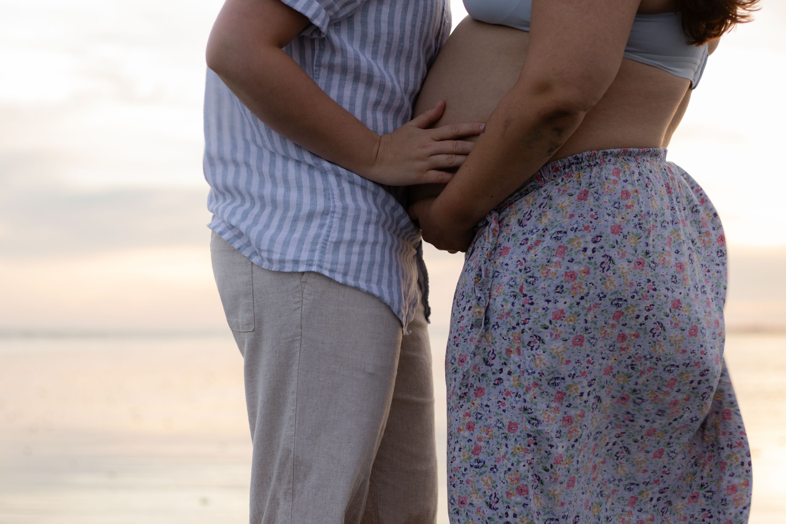 Couple tenderly holding pregnant belly on beach at sunset, wearing casual summer clothes.