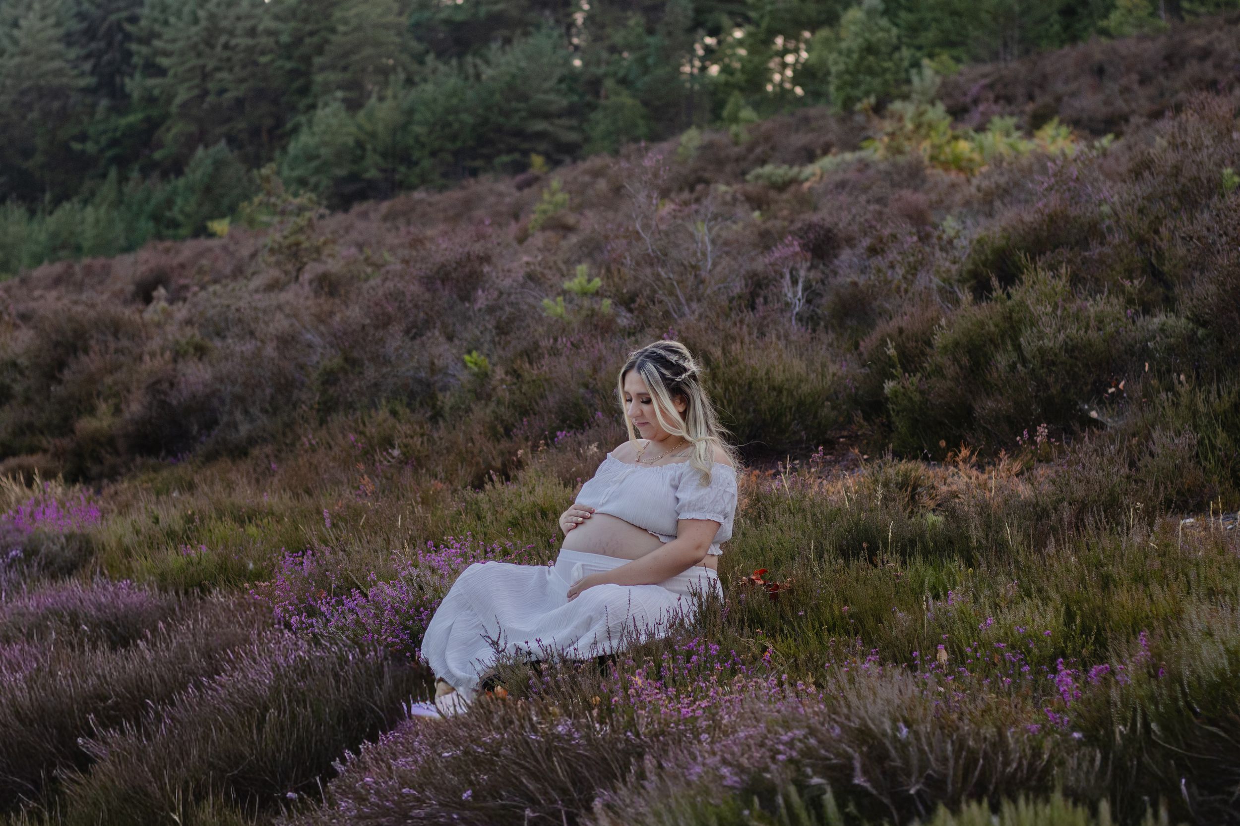 Pregnant woman in white dress sitting among purple heather flowers in a scenic outdoor field during golden hour.