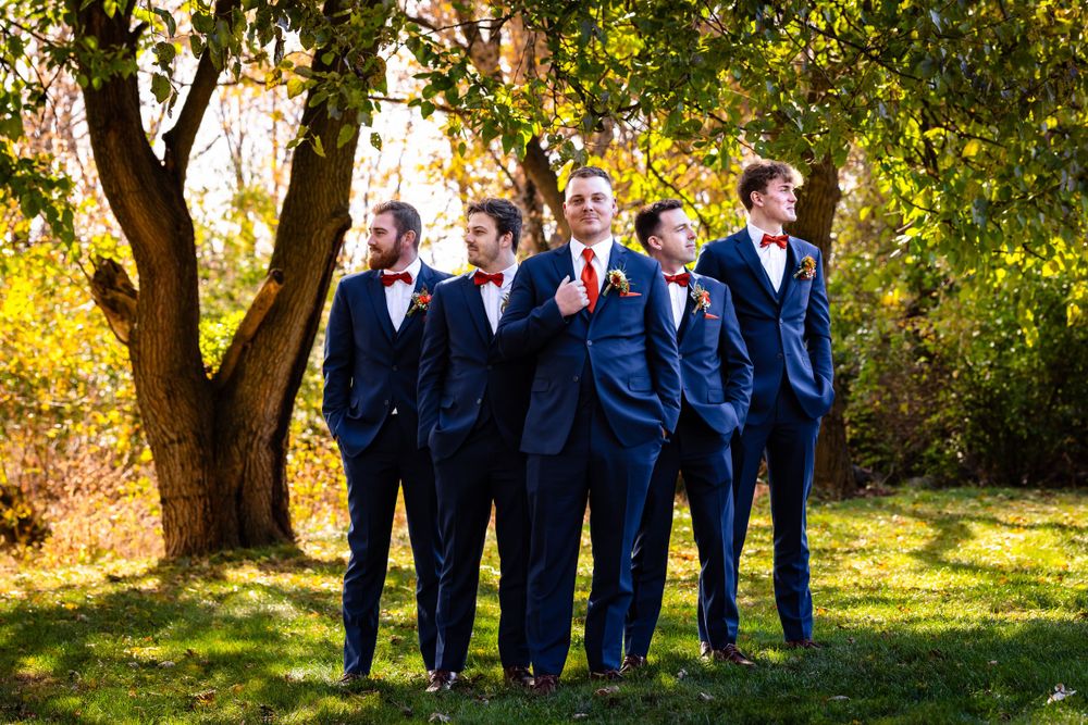 Wedding party in navy suits lined up for formal photo under autumn trees.