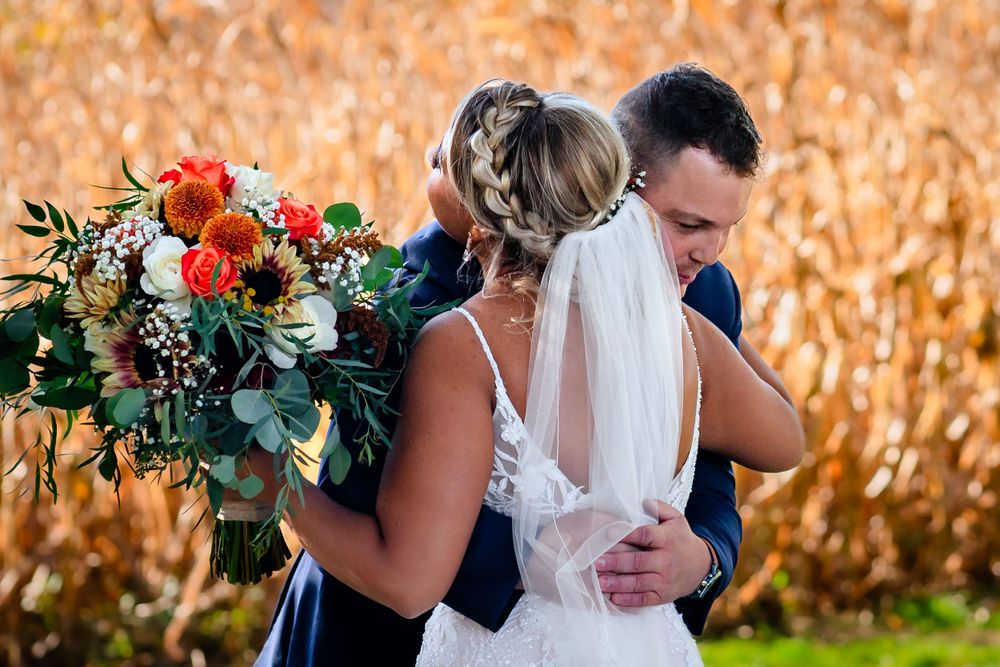 Wedding party poses playfully in front of autumn cornfield with orange and red floral bouquets.