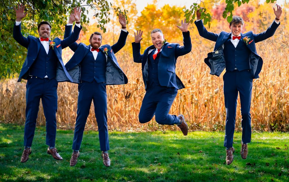 Wedding party jumps joyfully on grass with autumn foliage in background.