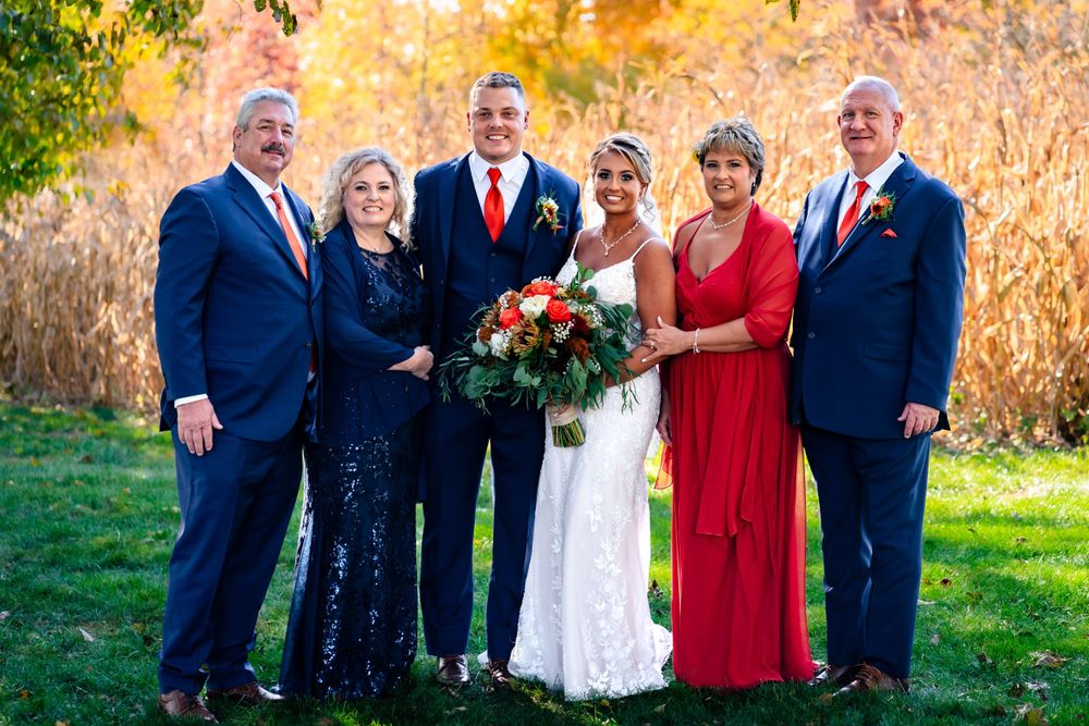 Wedding group poses cheerfully in mix of navy suits and red dresses against fall backdrop.