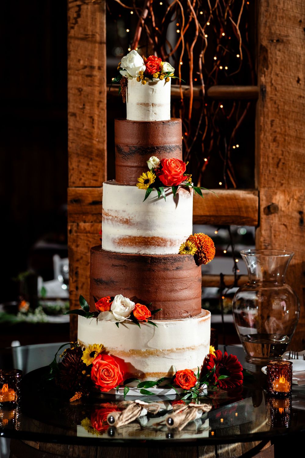 Semi-naked wedding cake decorated with fresh orange roses and flowers on a glass stand.