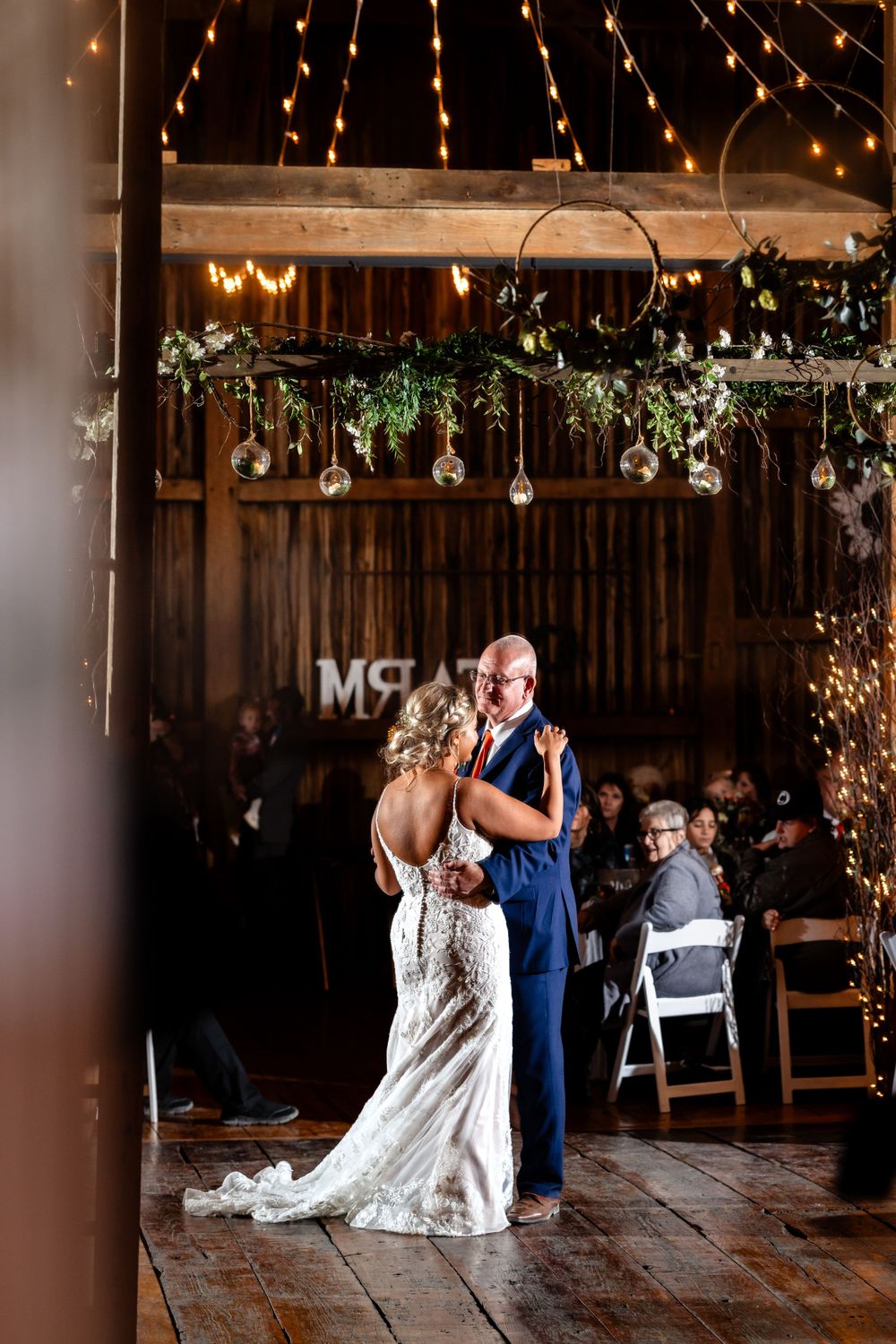 Couple dances beneath hanging greenery and string lights at rustic barn reception.