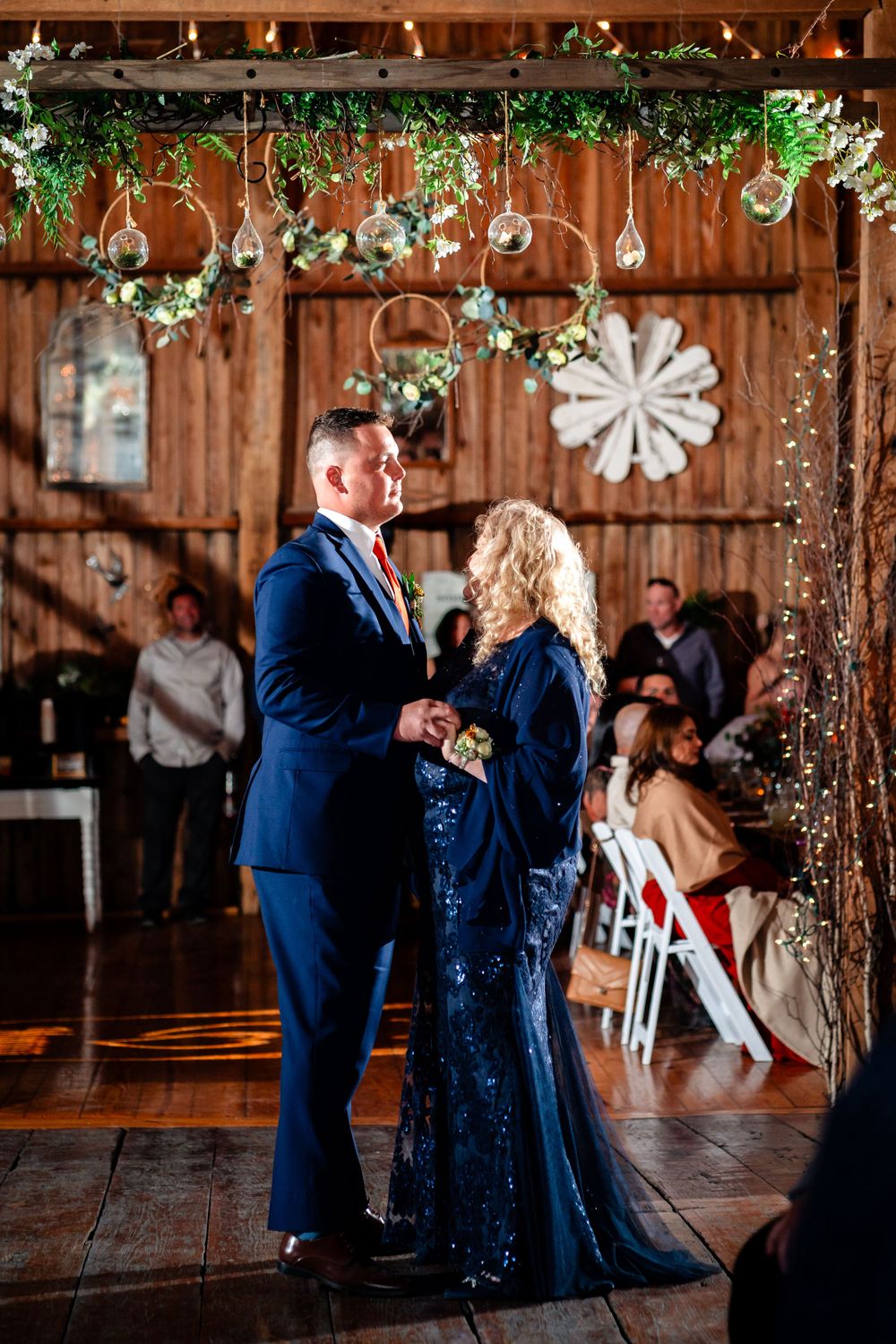 Couple dances beneath hanging botanical decorations in rustic barn setting.