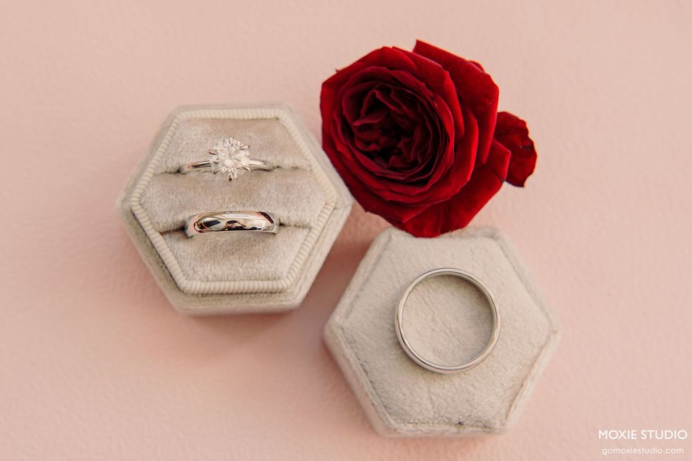 A silver engagement ring with a diamond solitaire sits elegantly beside a deep red rose on a soft pink background.