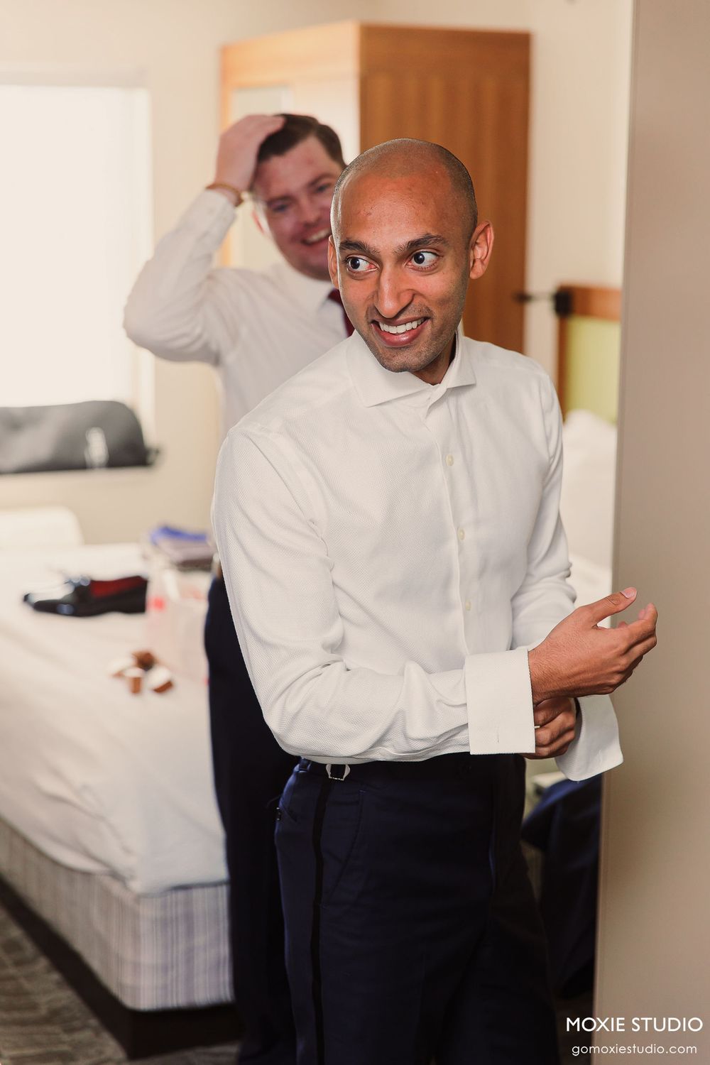 Groom getting ready for wedding ceremony in hotel room wearing white dress shirt and black pants.