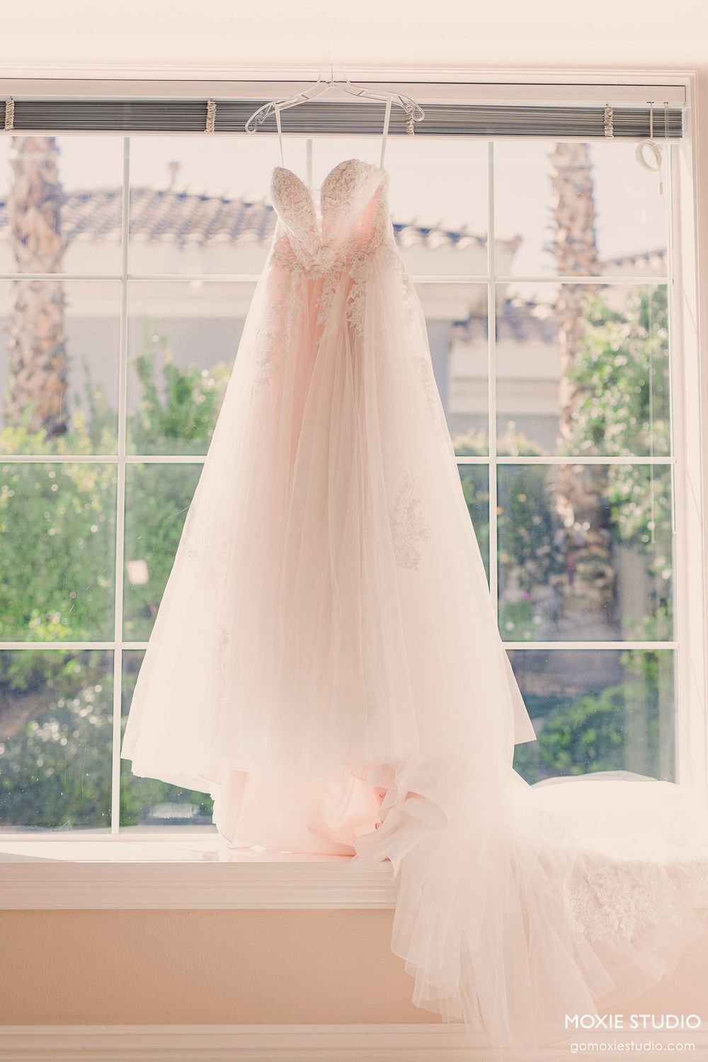 A white wedding dress hangs elegantly in front of sunlit windows in a bridal preparation room.
