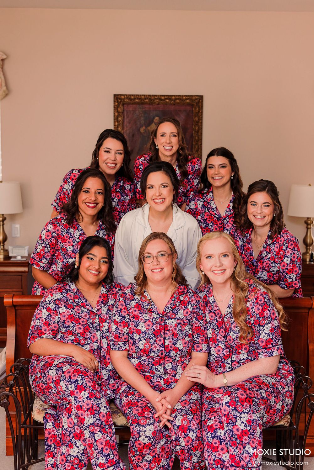 A bridal party wearing matching floral pink robes gather for a pre-wedding photo in a bedroom.
