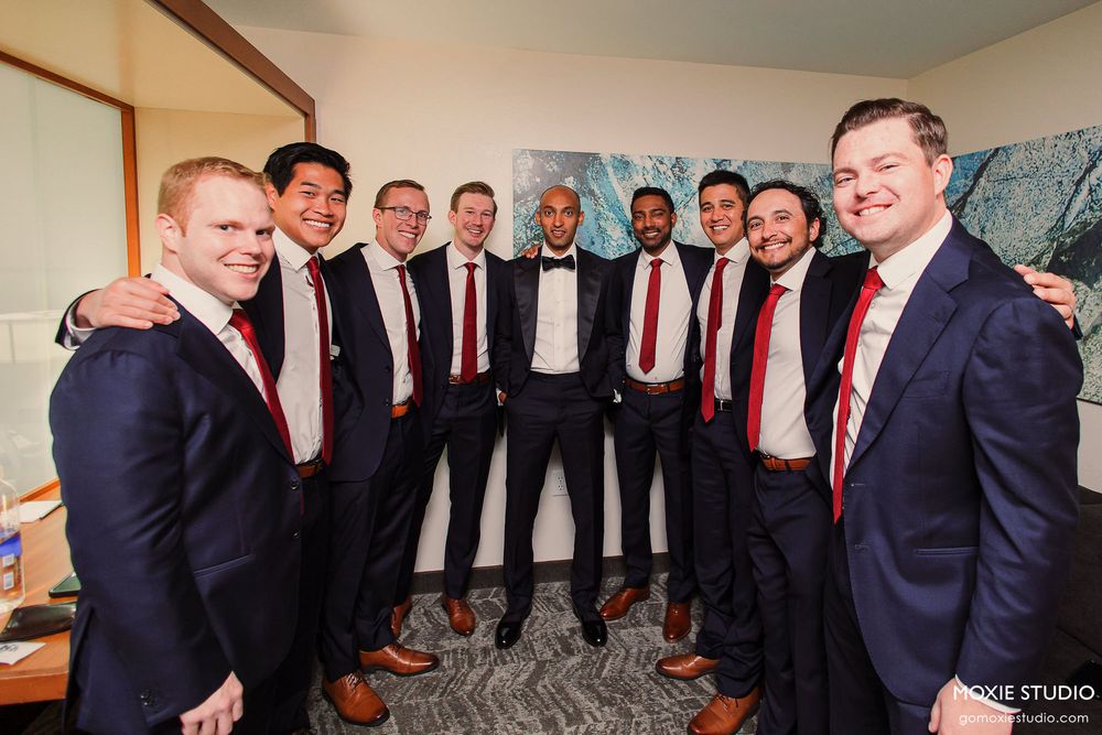 A wedding party group of groomsmen in navy suits with red ties pose together in a room.