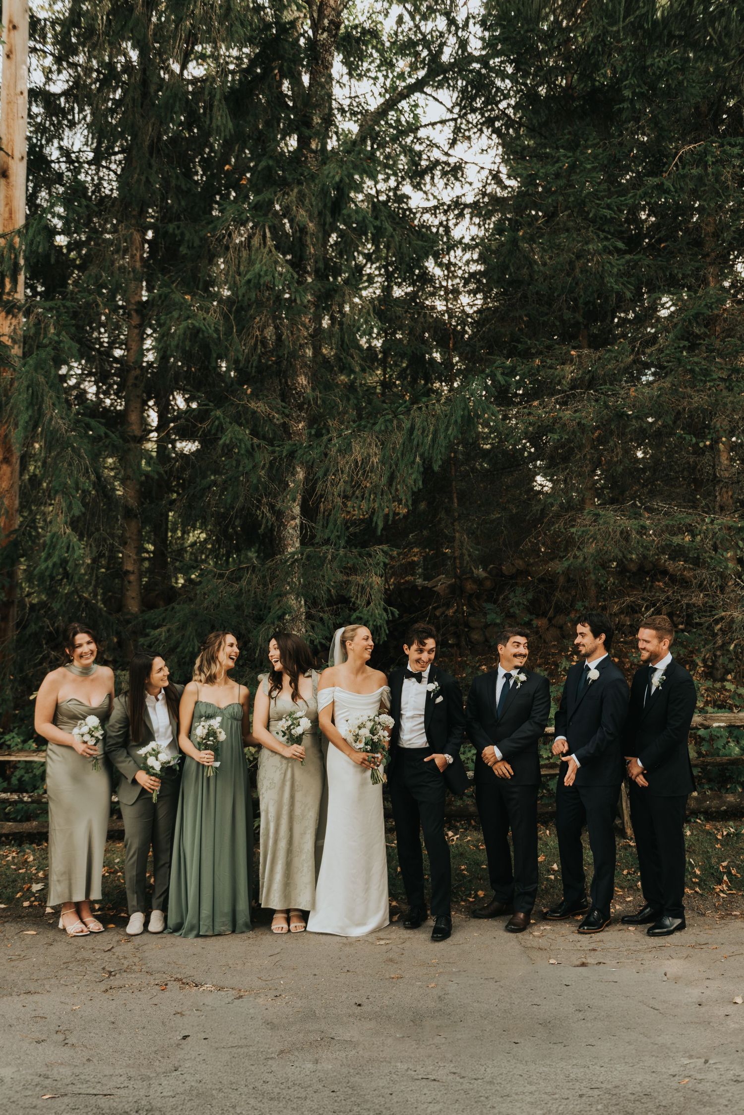 Wedding party in sage green dresses and black suits stands in line against wooded backdrop at outdoor ceremony.