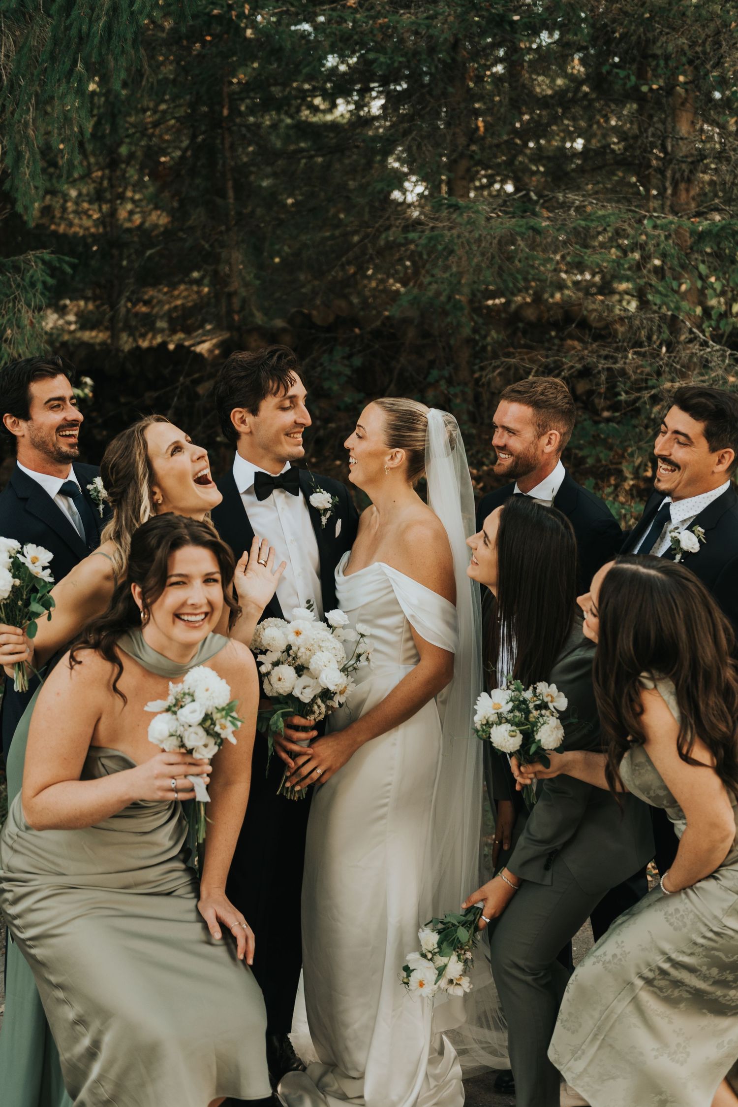 Wedding party laughs together in a candid moment outdoors with bridesmaids in sage dresses and white bouquets.
