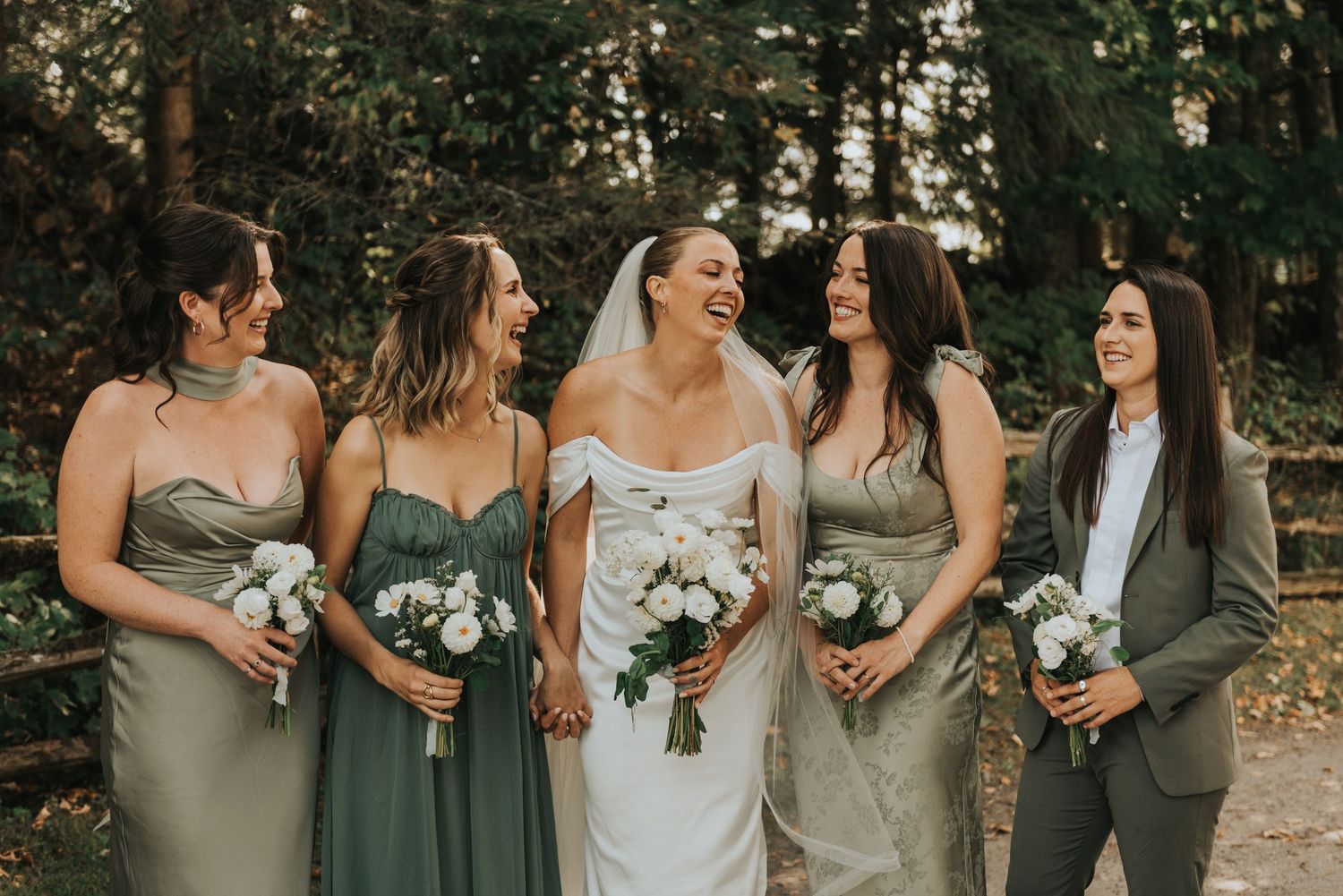 Wedding party in sage green dresses and gray suits pose together in a wooded outdoor setting.