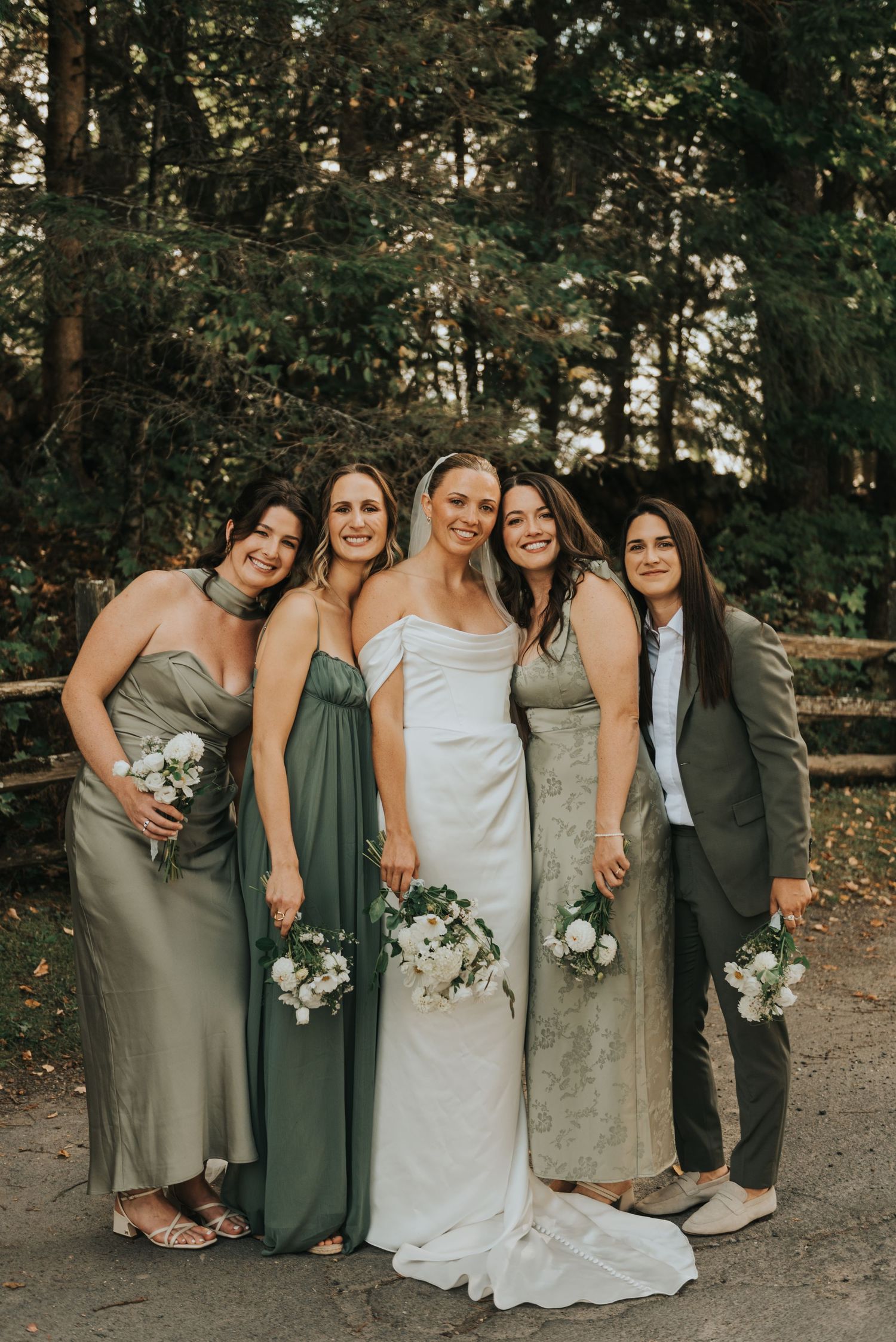 Wedding group poses together on forest path wearing sage green and white dresses with natural bouquets.