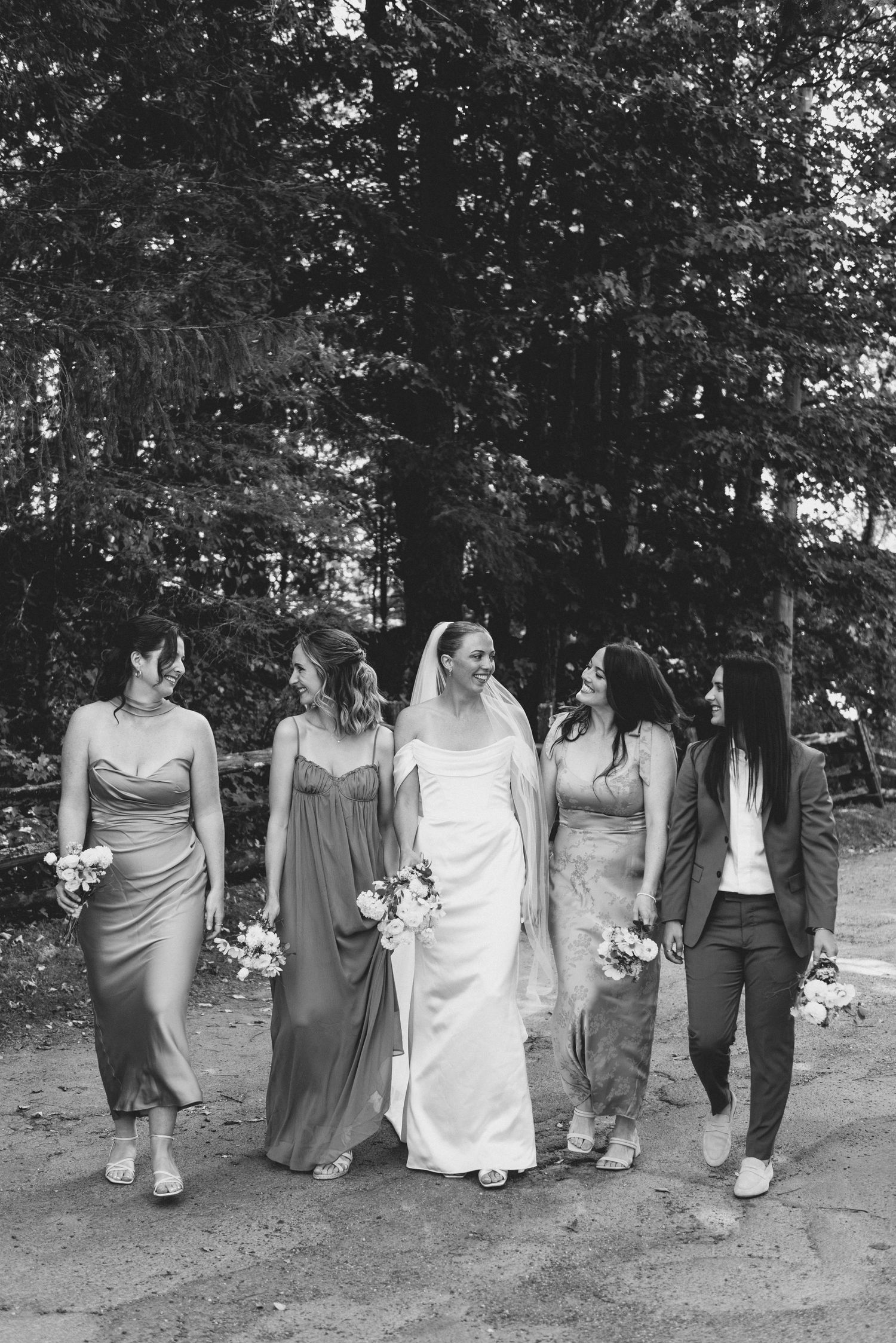 Wedding party walking in a line along a wooded path wearing gray dresses and carrying white bouquets.