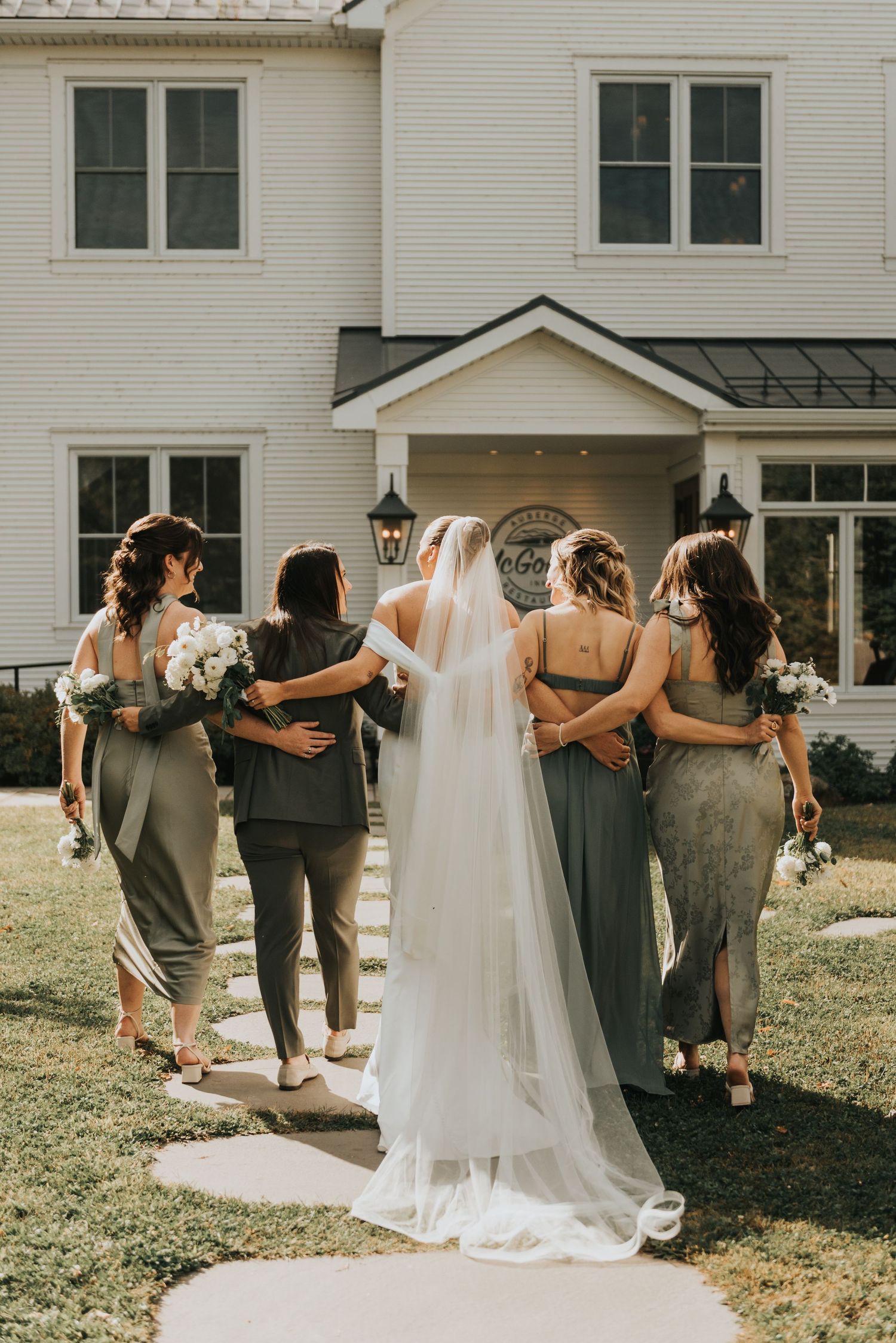 Bridal party in gray dresses poses with their backs to the camera against a white building backdrop.