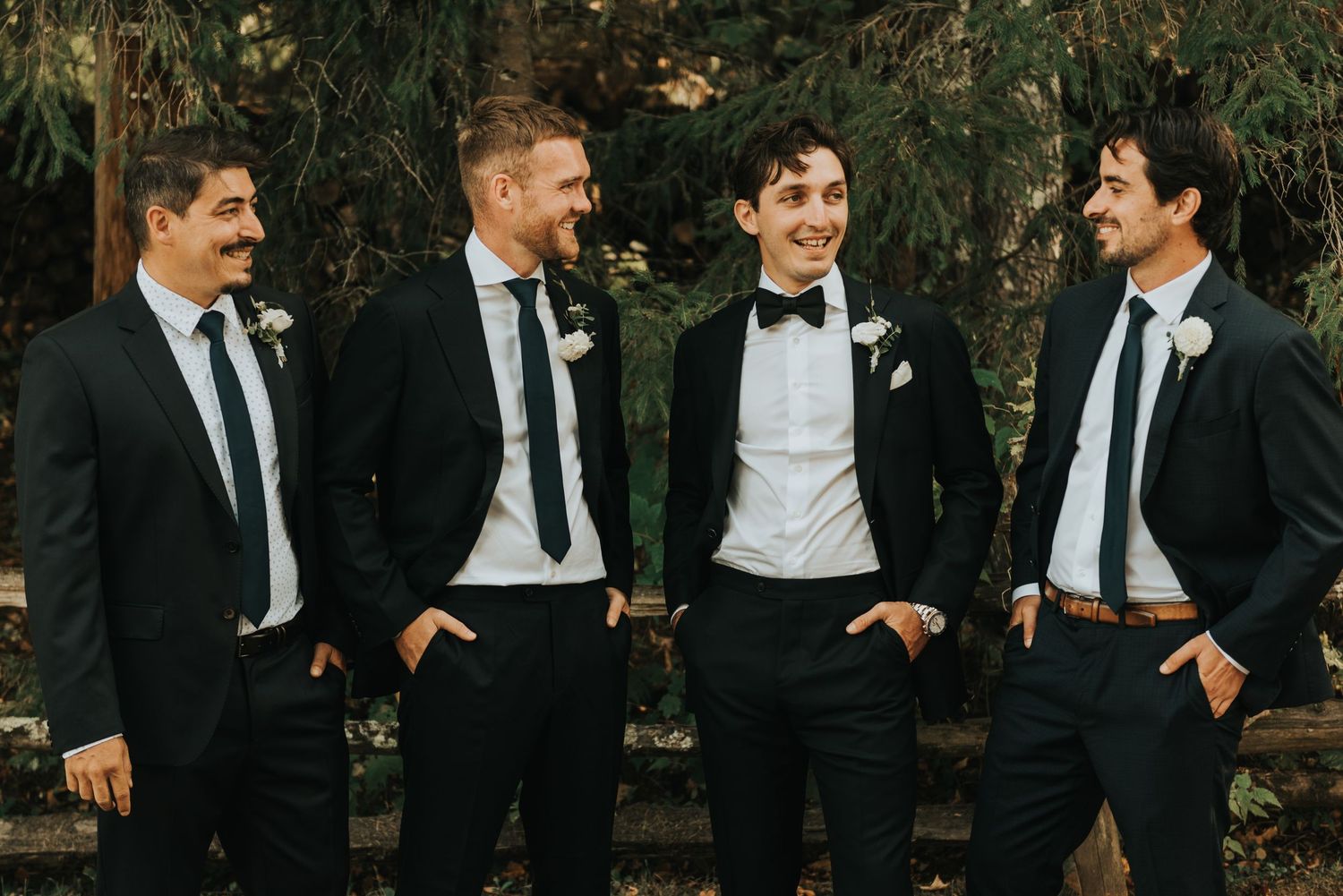 Groomsmen in black suits and ties share a candid laughing moment together outdoors during wedding festivities.