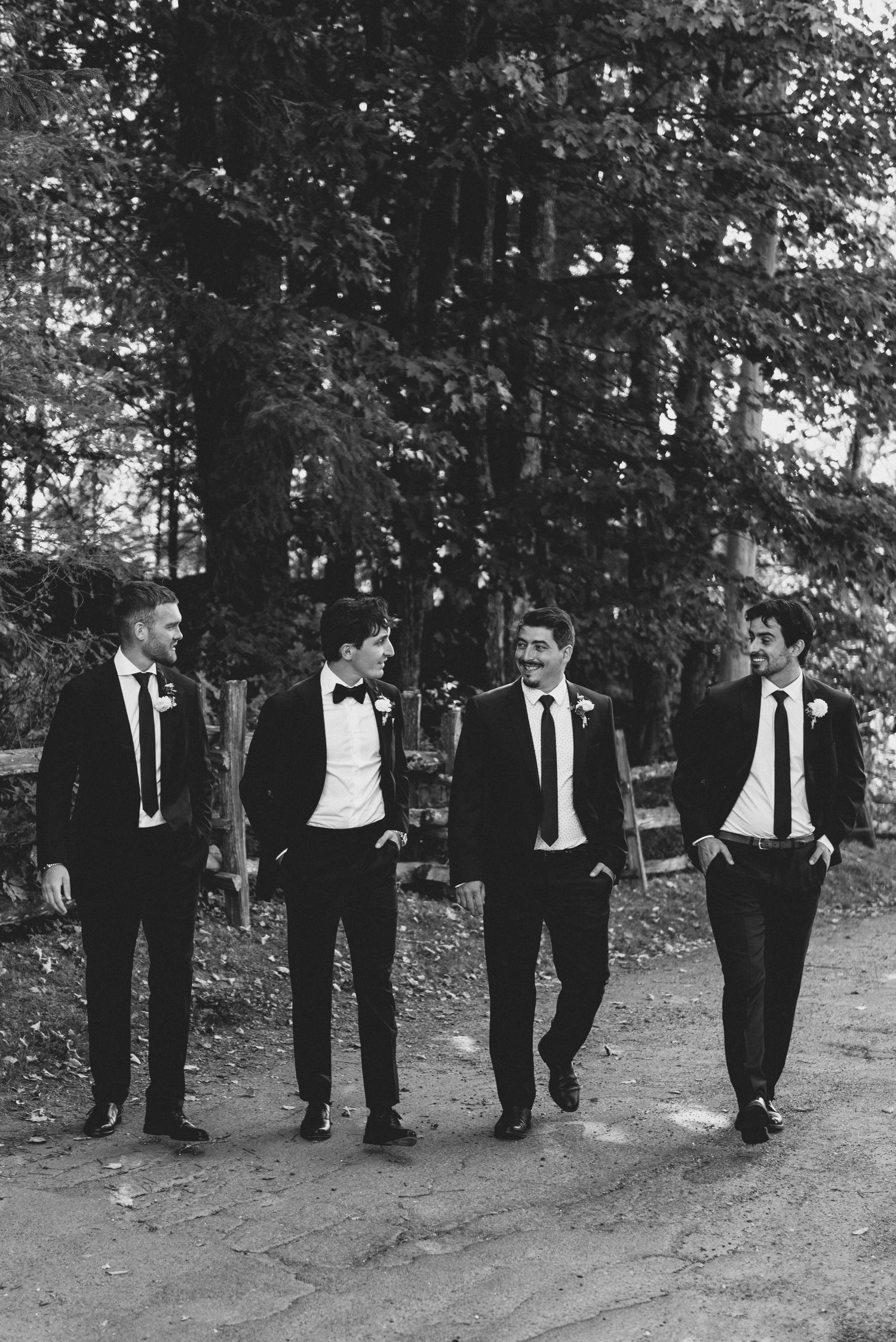 Groomsmen in black tuxedos walk in formation along an outdoor path during a wedding photo session.