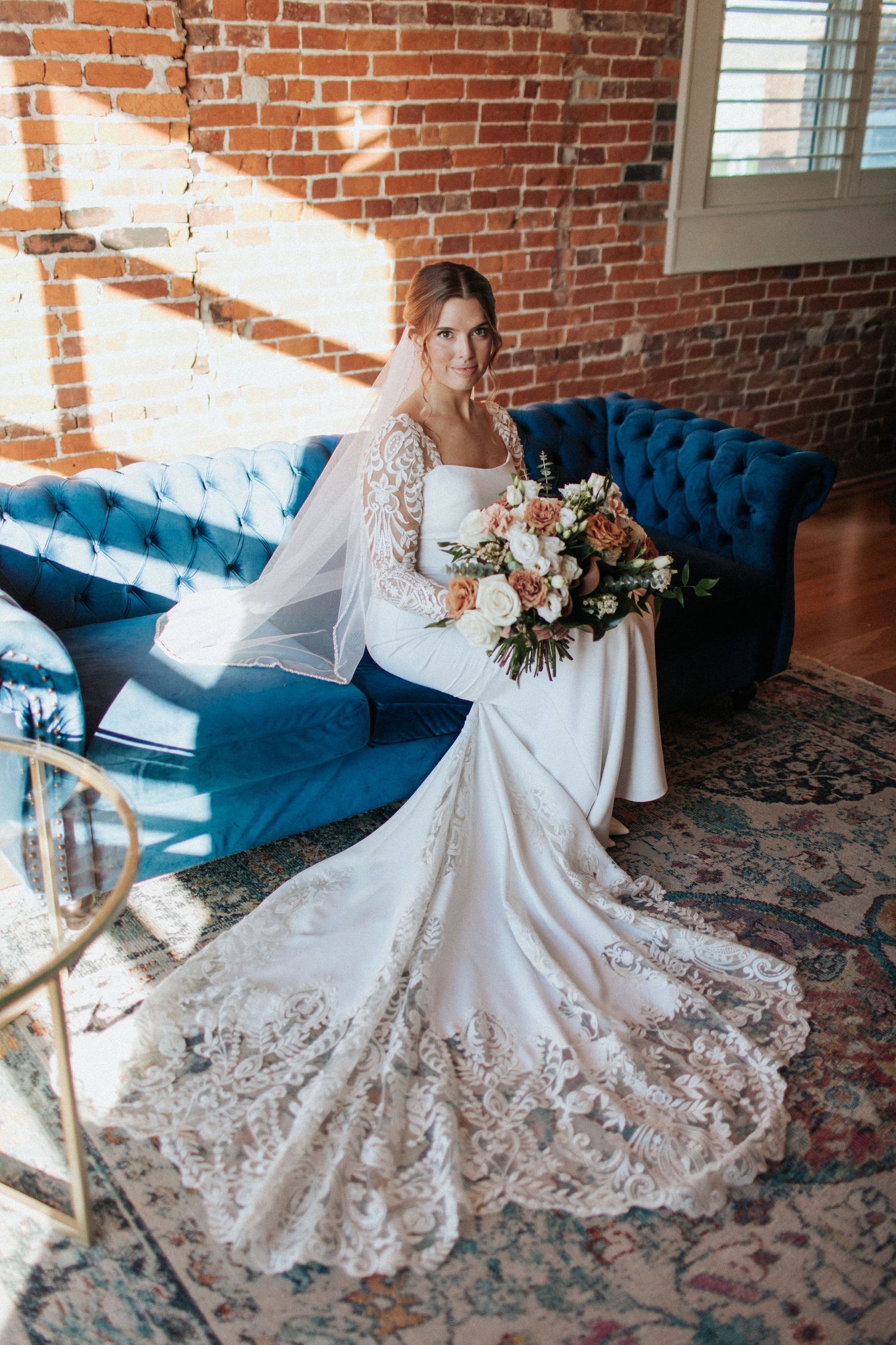 A wedding dress with delicate lace details and a flowing train displayed against a blue velvet couch and brick wall backdrop.