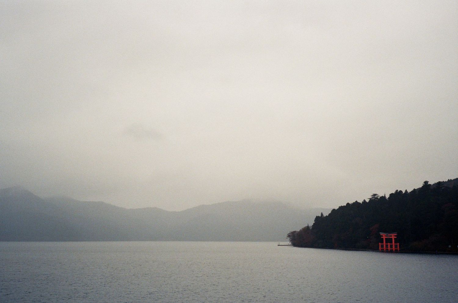 Misty panoramic view of a serene lake surrounded by mountains and forests with a traditional red torii gate in the distance.