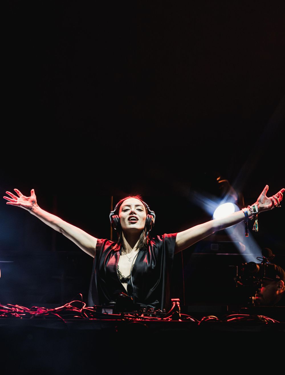 DJ performing at a nightclub with arms raised in celebration while mixing music behind decks in dramatic lighting.