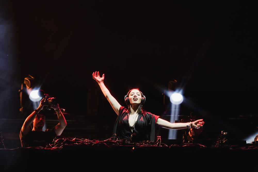 Dark concert stage with dramatic spotlights and raised hands silhouetted against the background.