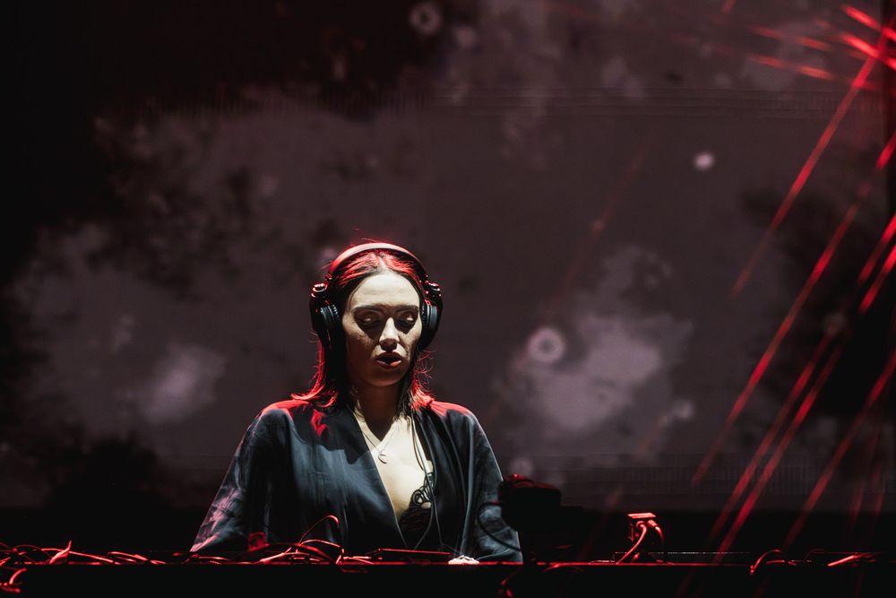 DJ performing at a dark nightclub with dramatic red lighting and electronic equipment.