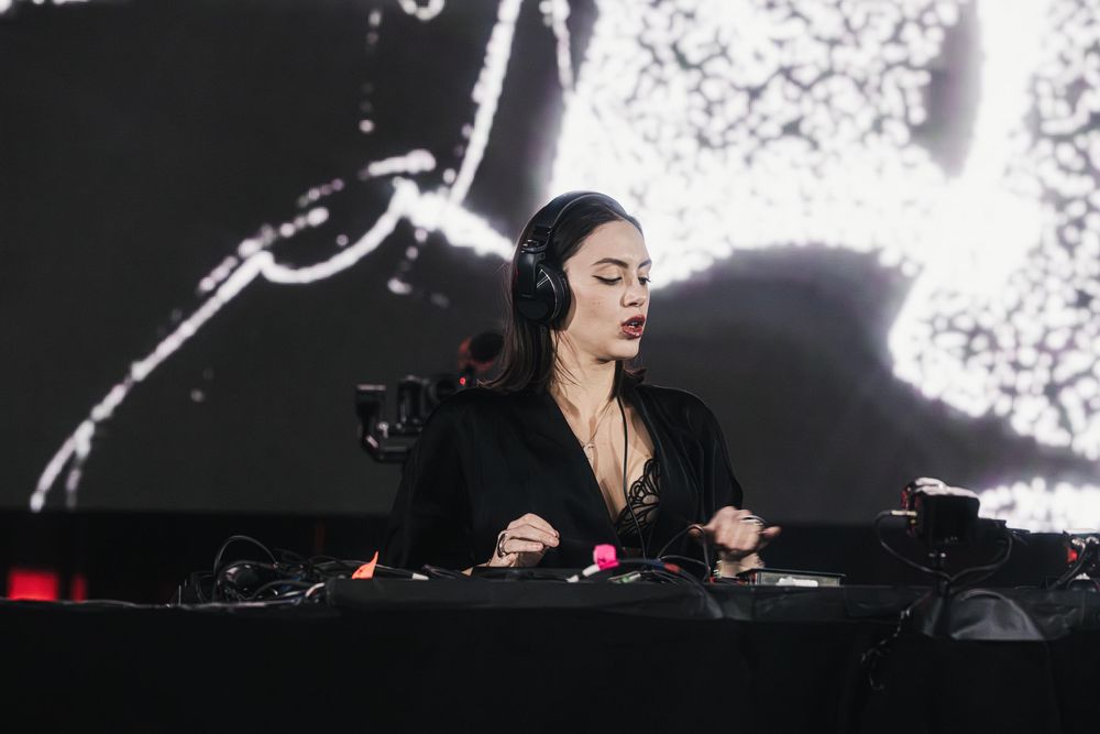 DJ performs behind decks at an electronic music event with dramatic lighting and visual effects in the background.